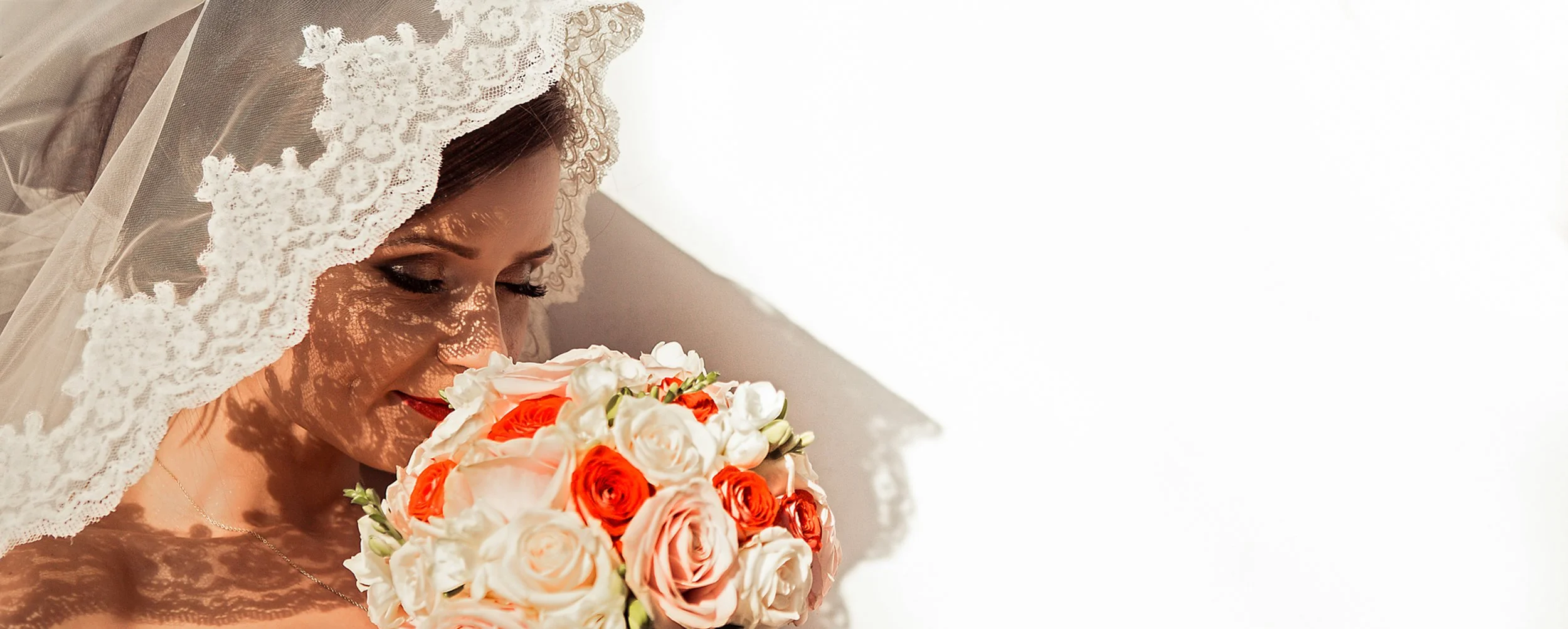 Close-up of a bride with lace veil holding a bouquet of white and peach roses, with shadows cast on her face.