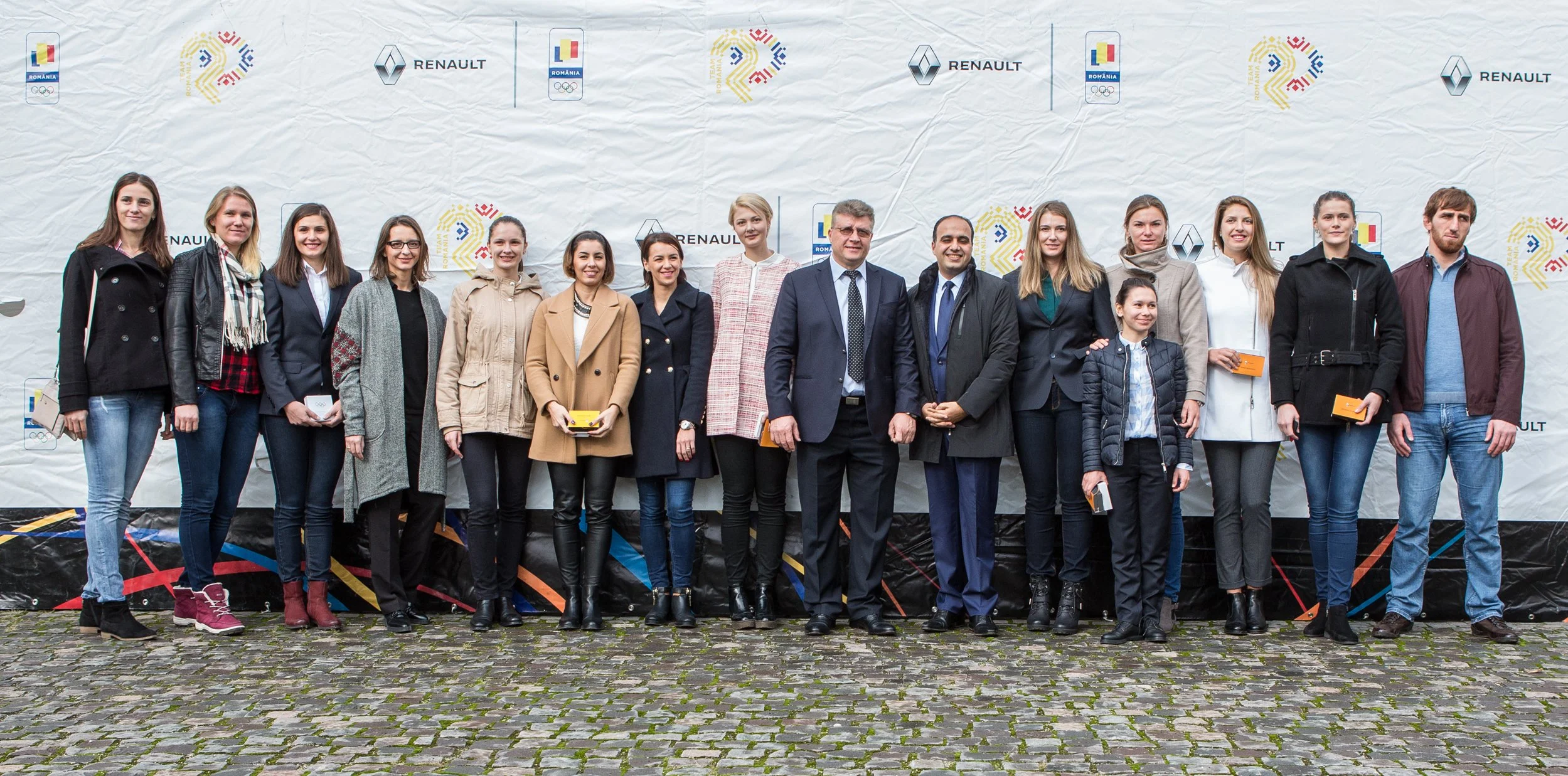 Group of people standing outdoors in front of a branded backdrop, some holding small boxes, smiling for a photo.