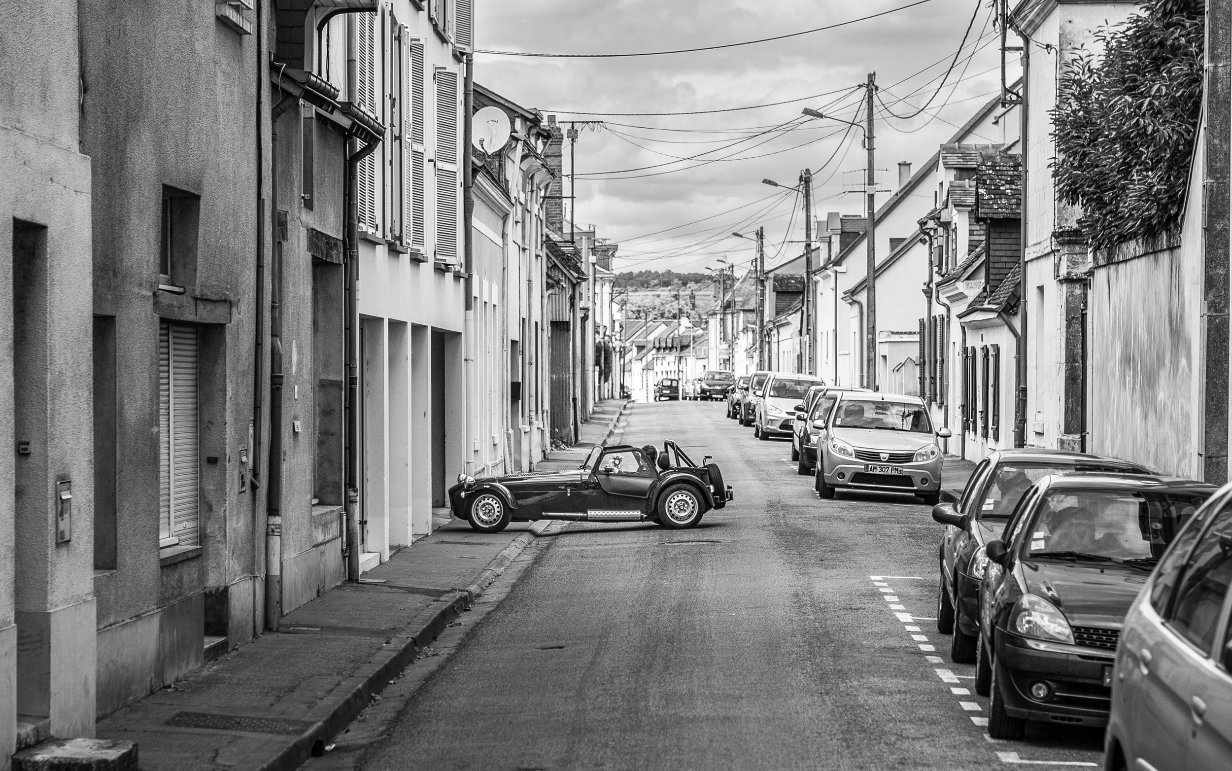 A black-and-white photo of a quiet residential street with parked cars lining the curb and an old-fashioned mini convertible car parked on the sidewalk. The buildings have various window styles and electrical wires crisscross above, with a cloudy sky