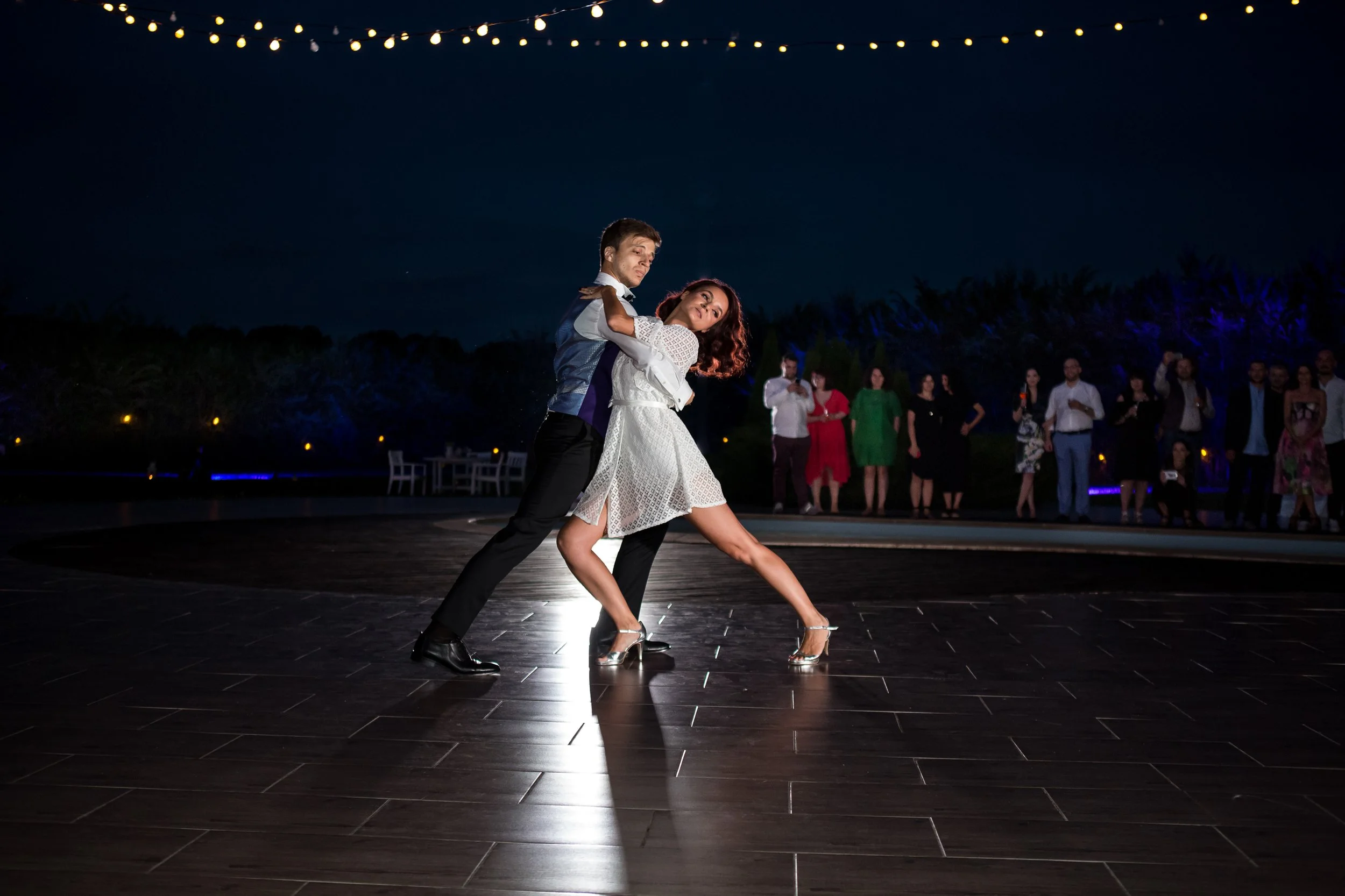 Couple dancing at night outdoors, surrounded by onlookers under string lights.
