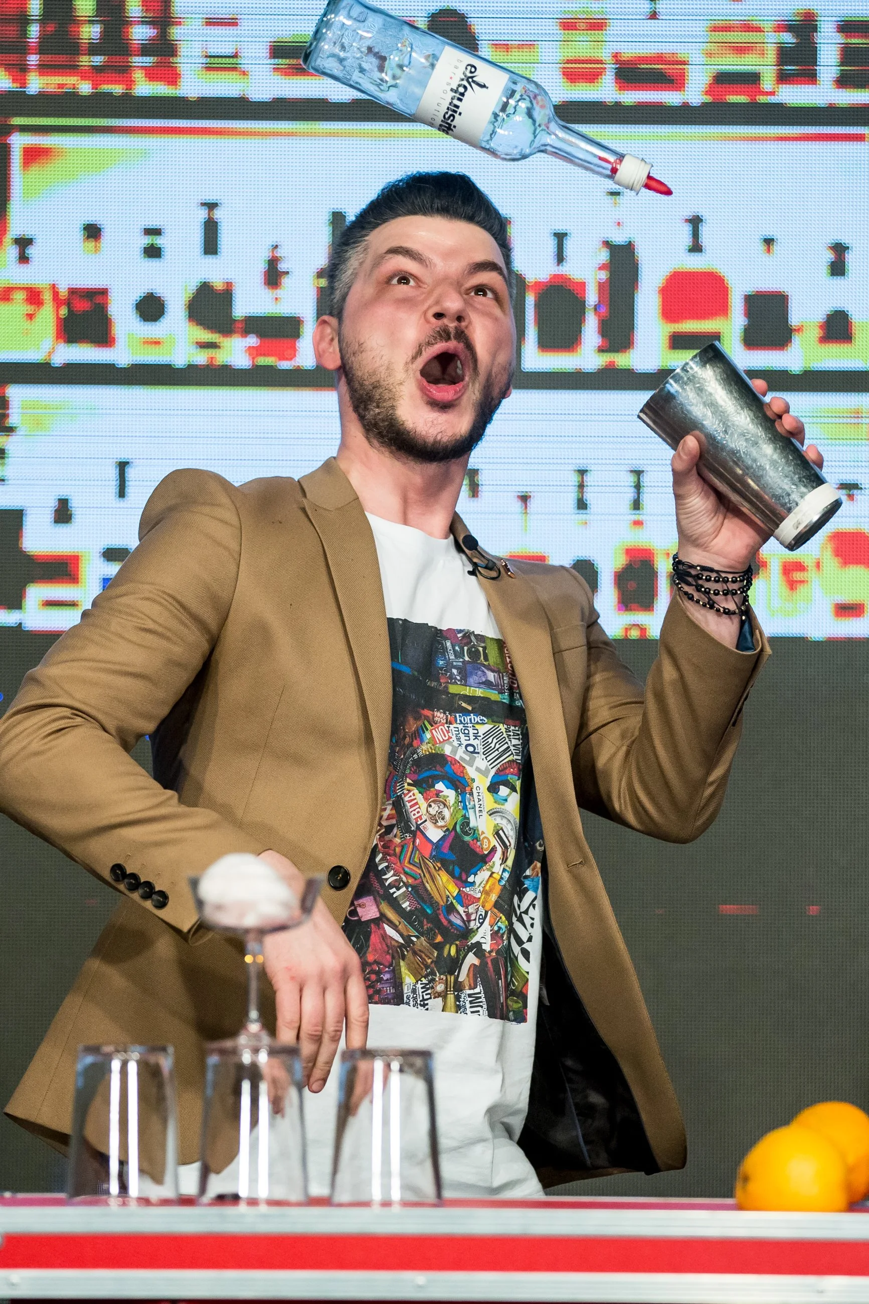 Man in a tan blazer and graphic T-shirt throwing a shot glass into the air at a table with glasses and lemons.