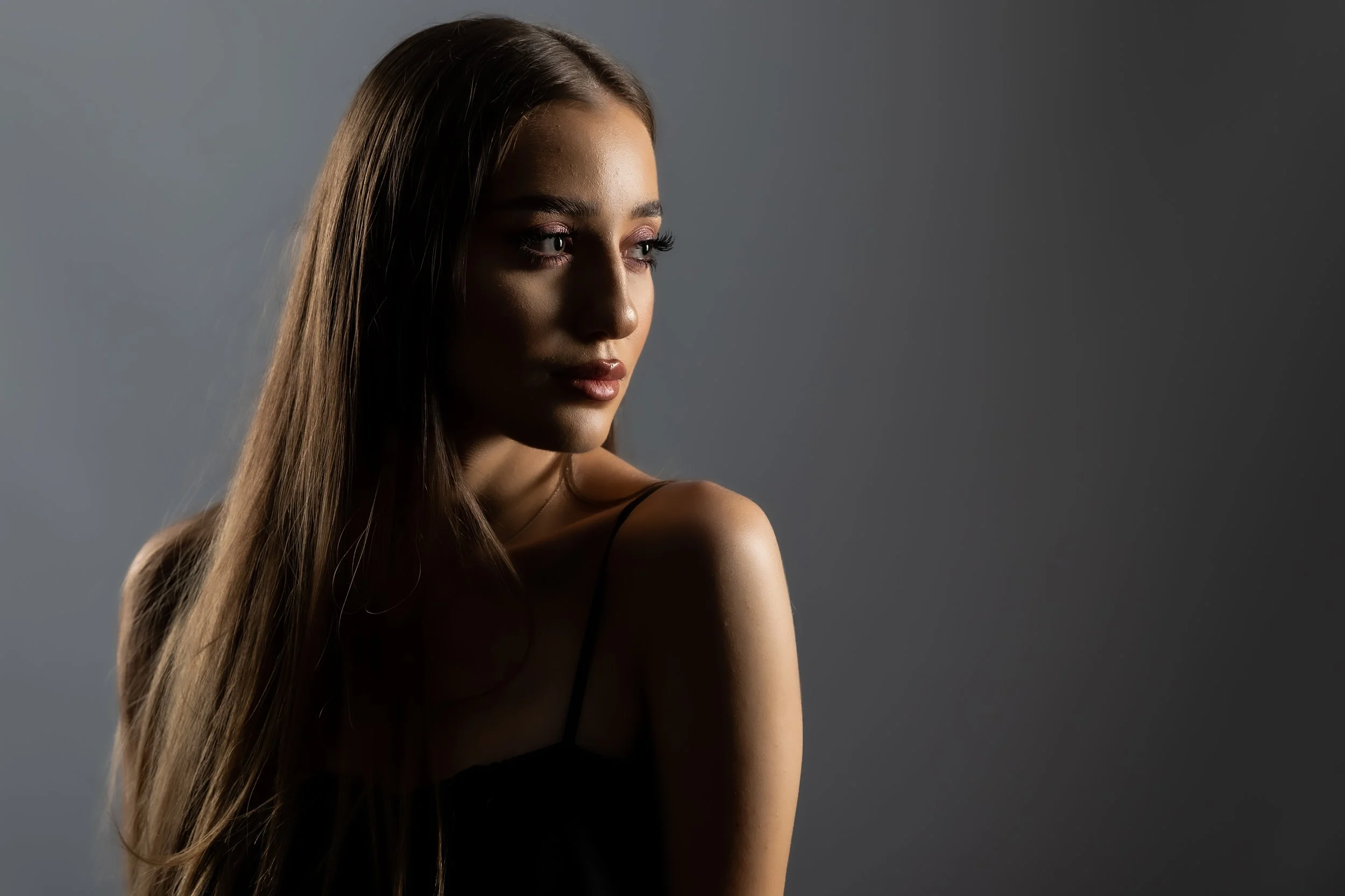 A young woman with long, straight brown hair looking to her left against a dark gray background.