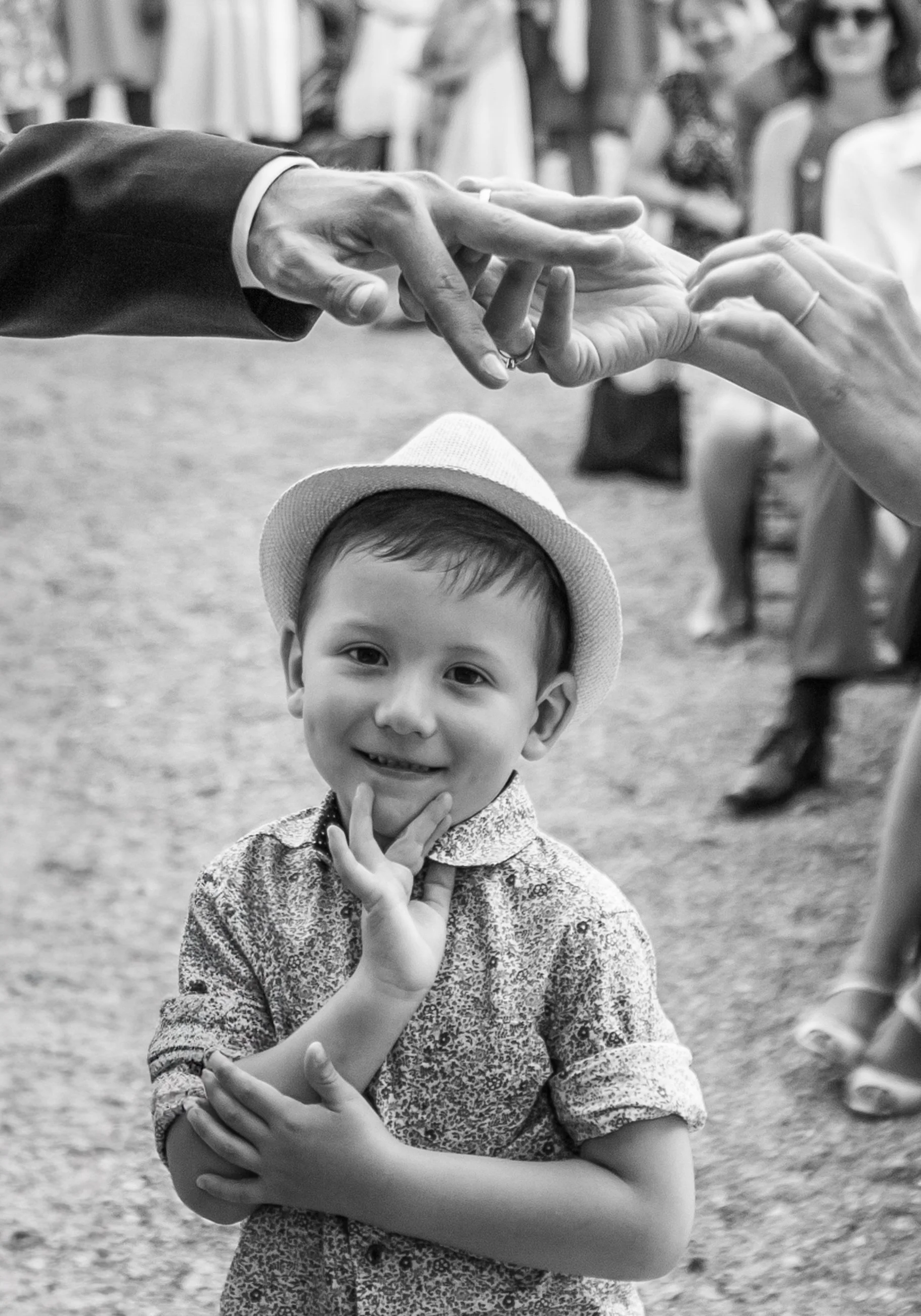 A young boy with a smile and a finger on his chin, wearing a patterned shirt and a hat, standing outdoors among adults who are blurred in the background.