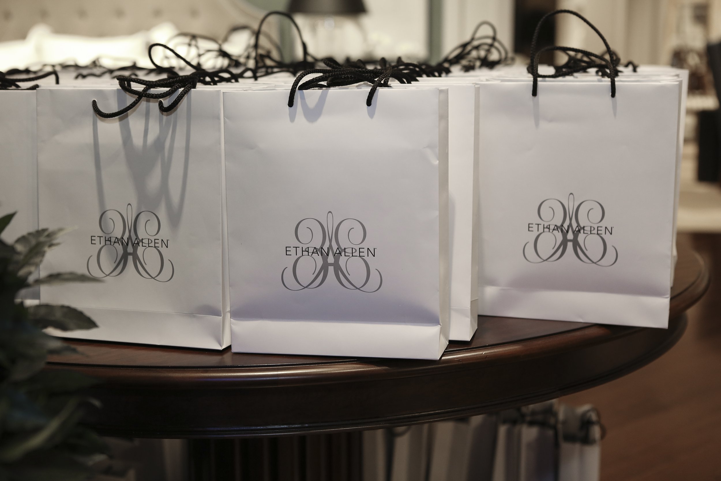 White gift bags with black string handles and the label 'Ethan Allen' featuring an intricate black swirl design, arranged on a dark wooden surface.