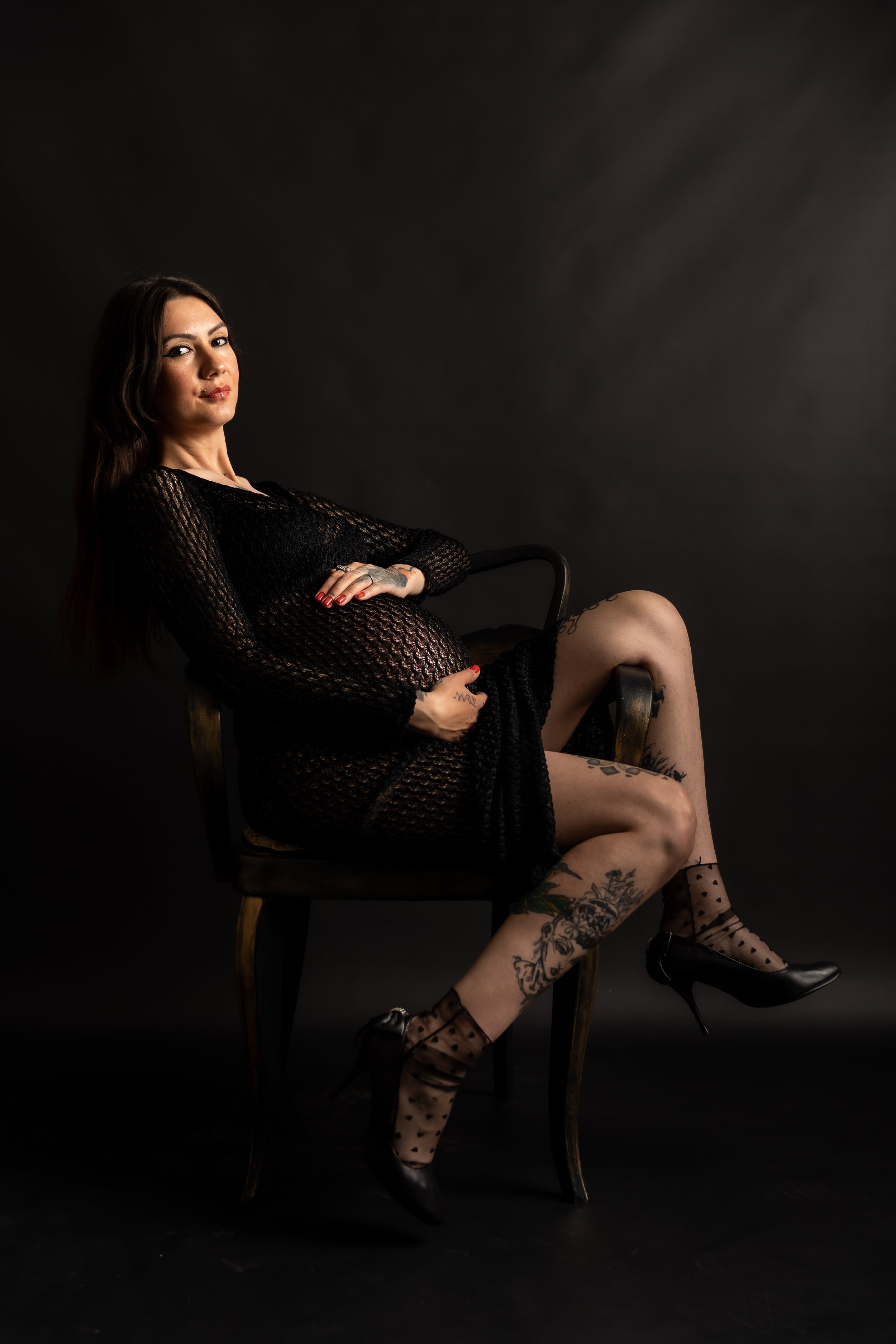 A woman with long brown hair sitting on a wooden chair against a dark background. She is wearing a black mesh dress, sheer polka dot stockings, and black high heels. She has tattoos on her legs and fingers, and her arms are crossed over her stomach.