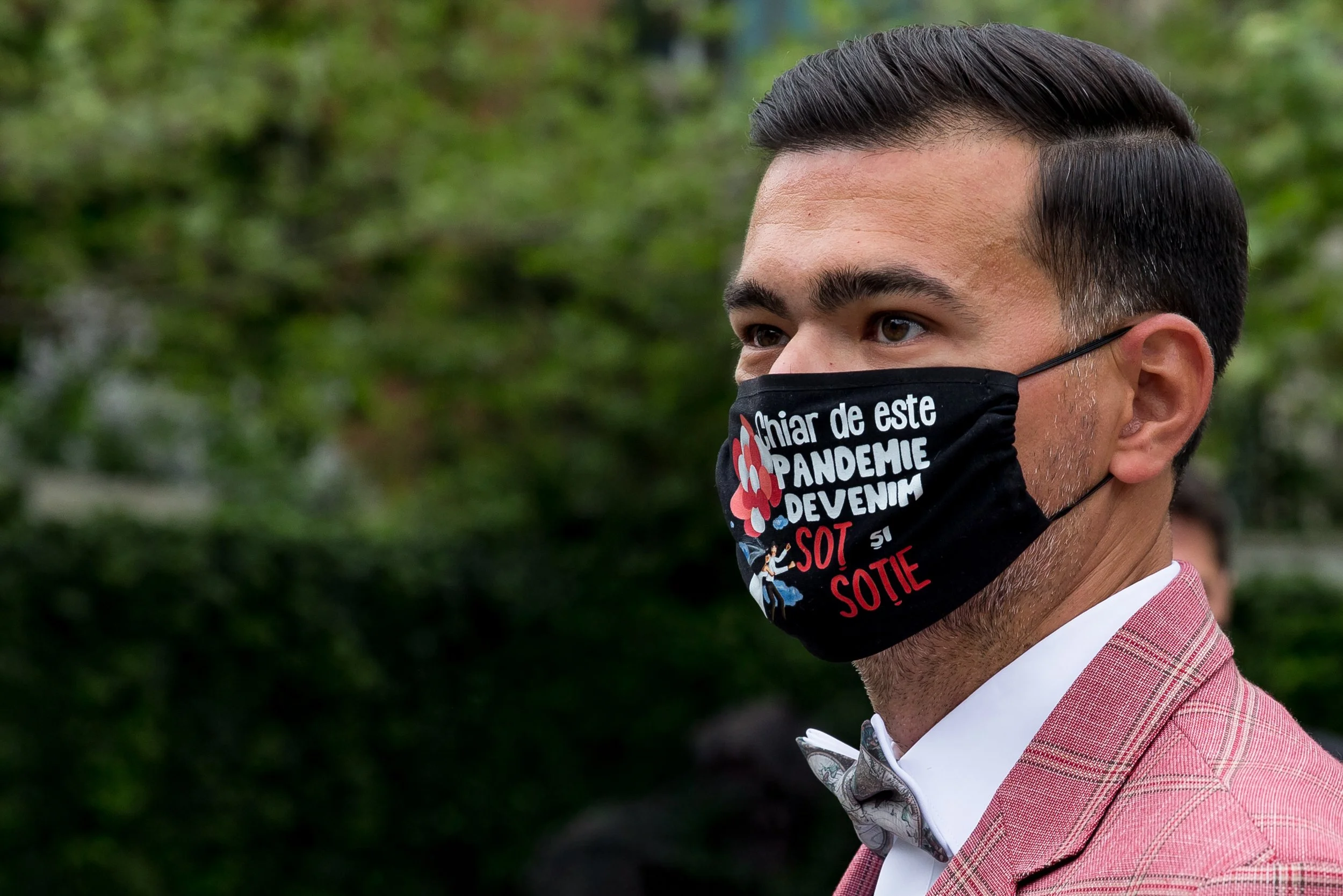 A man dressed in a pink suit with a white shirt and bow tie, wearing a black face mask with red and white text, standing outdoors with green foliage in the background.