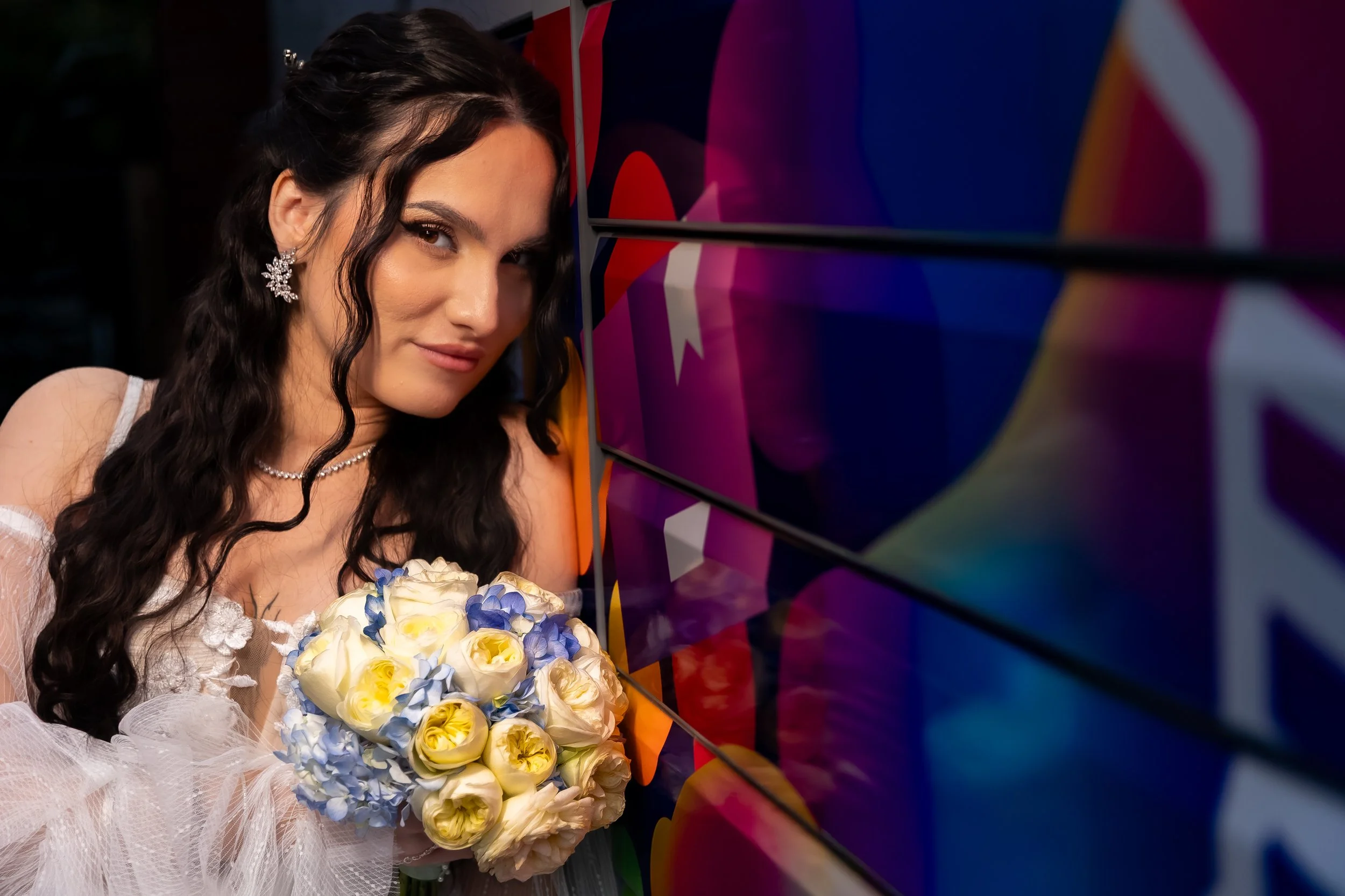 A woman with long dark curly hair wearing a white dress and earrings, holding a bouquet of white and yellow roses with blue accents, standing next to a colorful geometric mural.