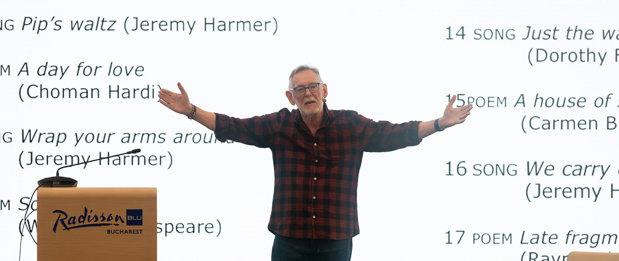 A man with glasses and gray hair wearing a plaid shirt is standing with his arms outstretched in front of a screen displaying a list of song titles and authors. There is a podium with a Radisson hotel logo in front of him.