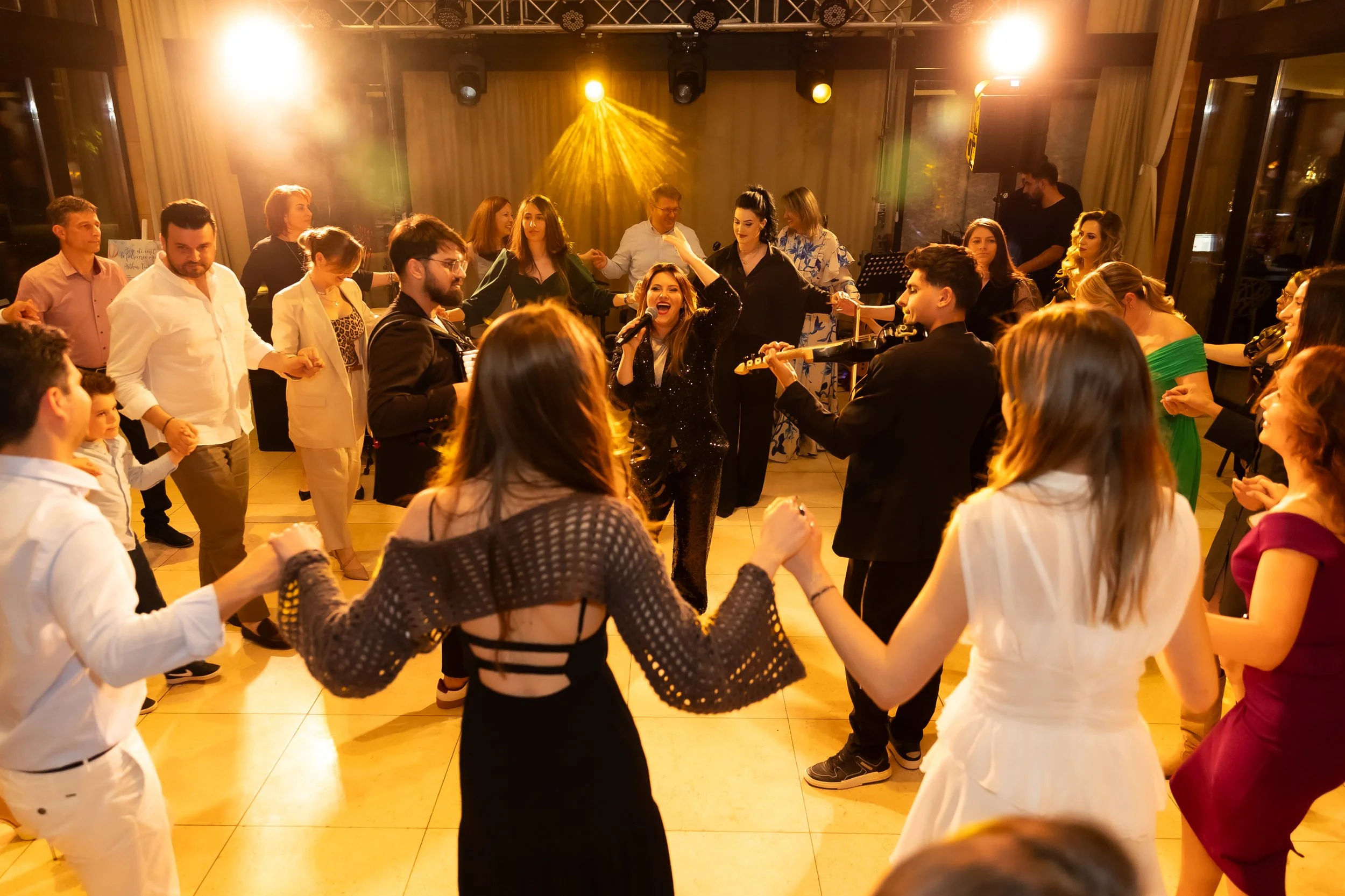 People dancing and singing at a party or celebration, with a woman in the center holding a microphone and smiling, surrounded by others holding hands and enjoying the moment, in a warmly lit indoor space.