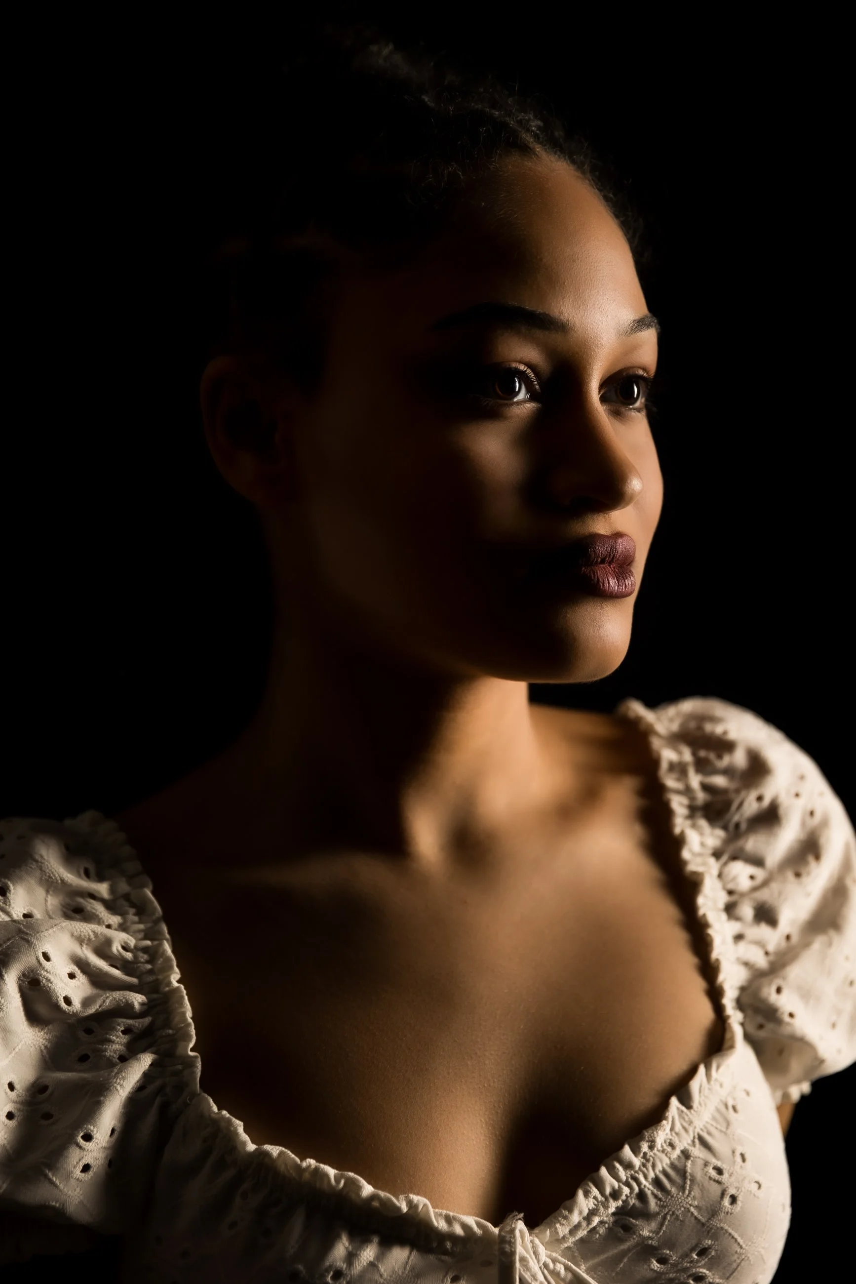 Close-up of a woman's face, partially illuminated with soft lighting, wearing a white eyelet blouse with puffed sleeves.