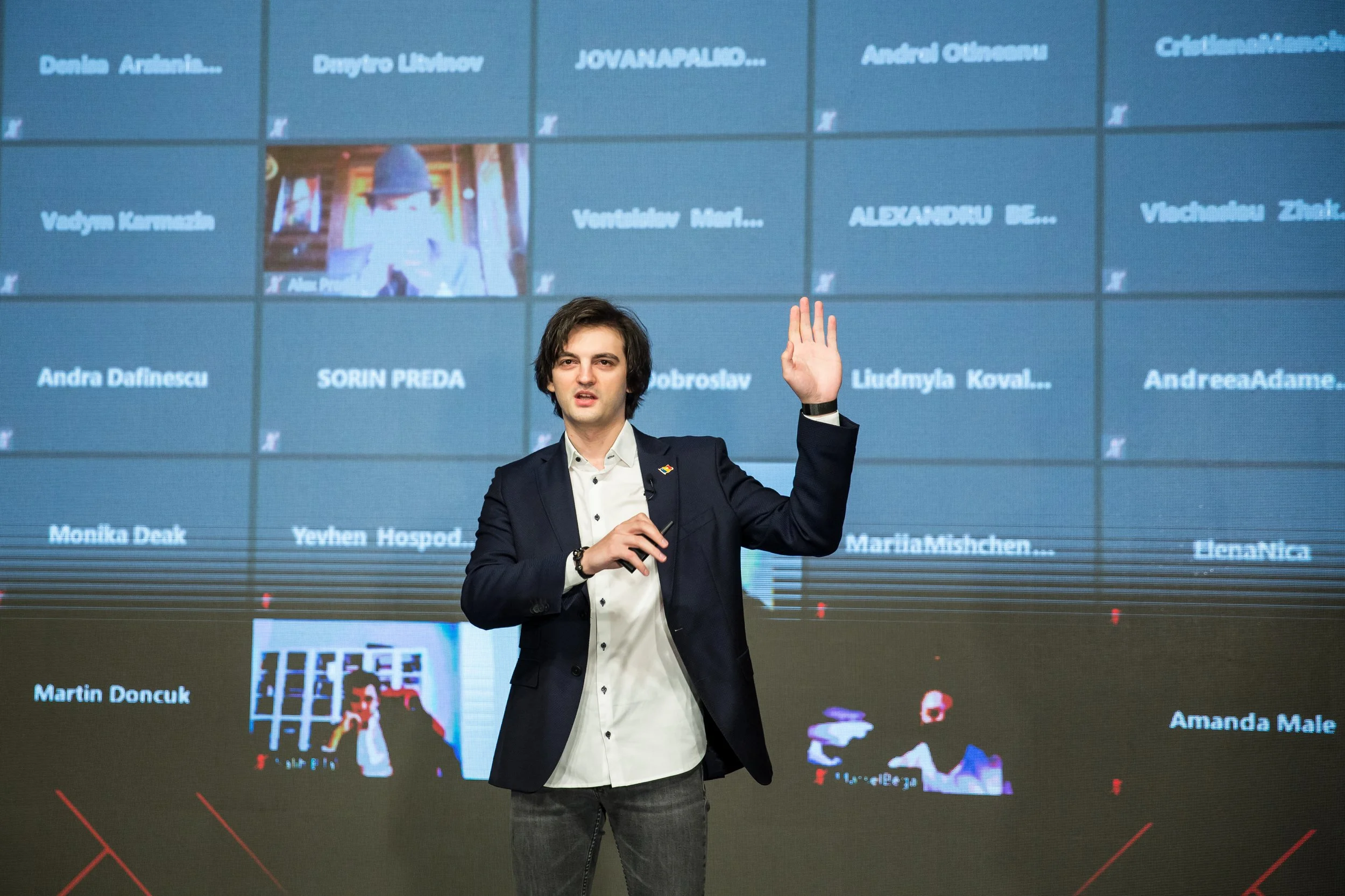 A young man in a suit with dark hair standing in front of a large screen displaying multiple names and images. He is raising one hand and appears to be speaking or presenting.