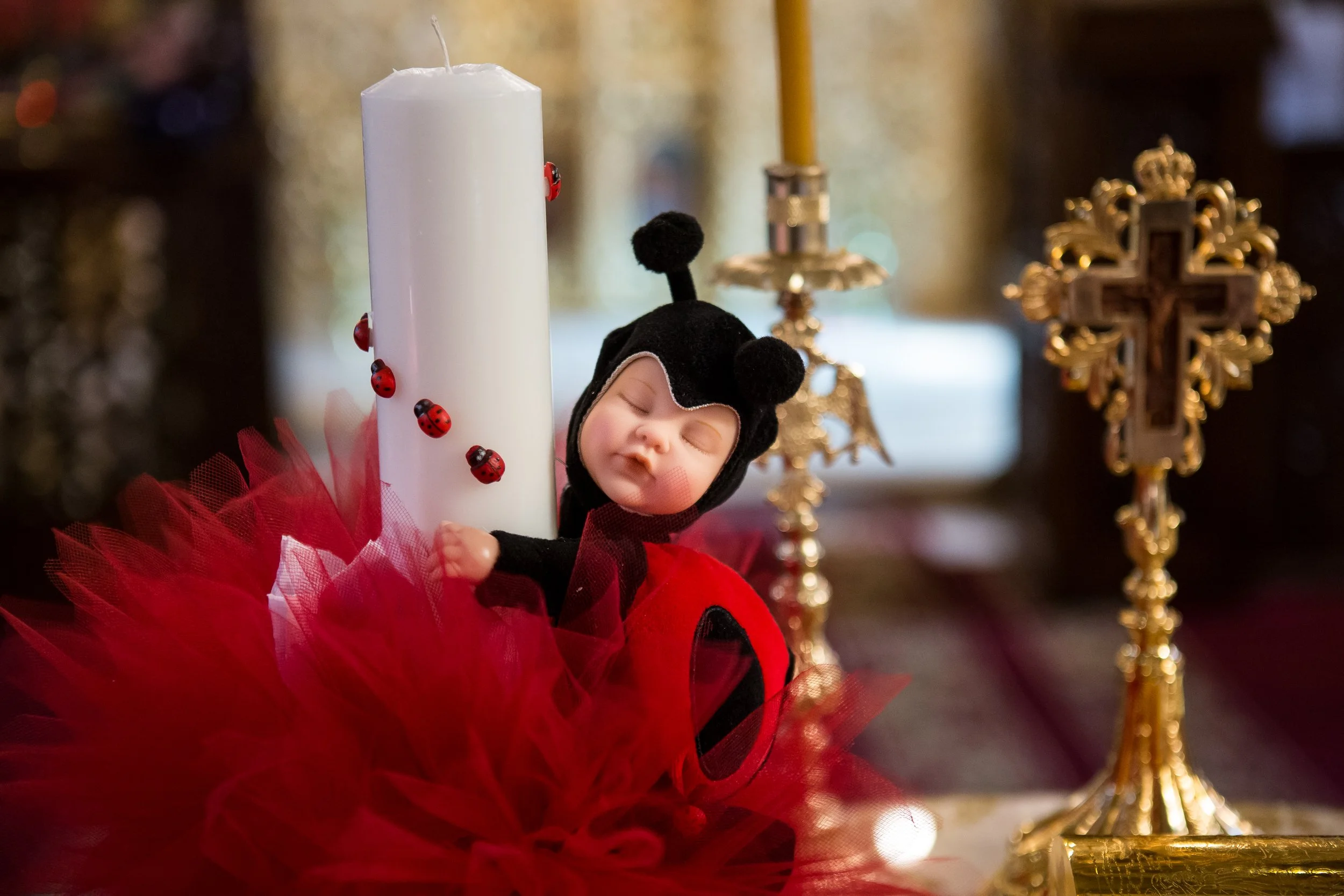 A small doll dressed in a ladybug costume with a black headband coral antennae, surrounded by a red tutu, placed in front of a white candle with ladybug decorations, and a gold Christmas ornament in the background.
