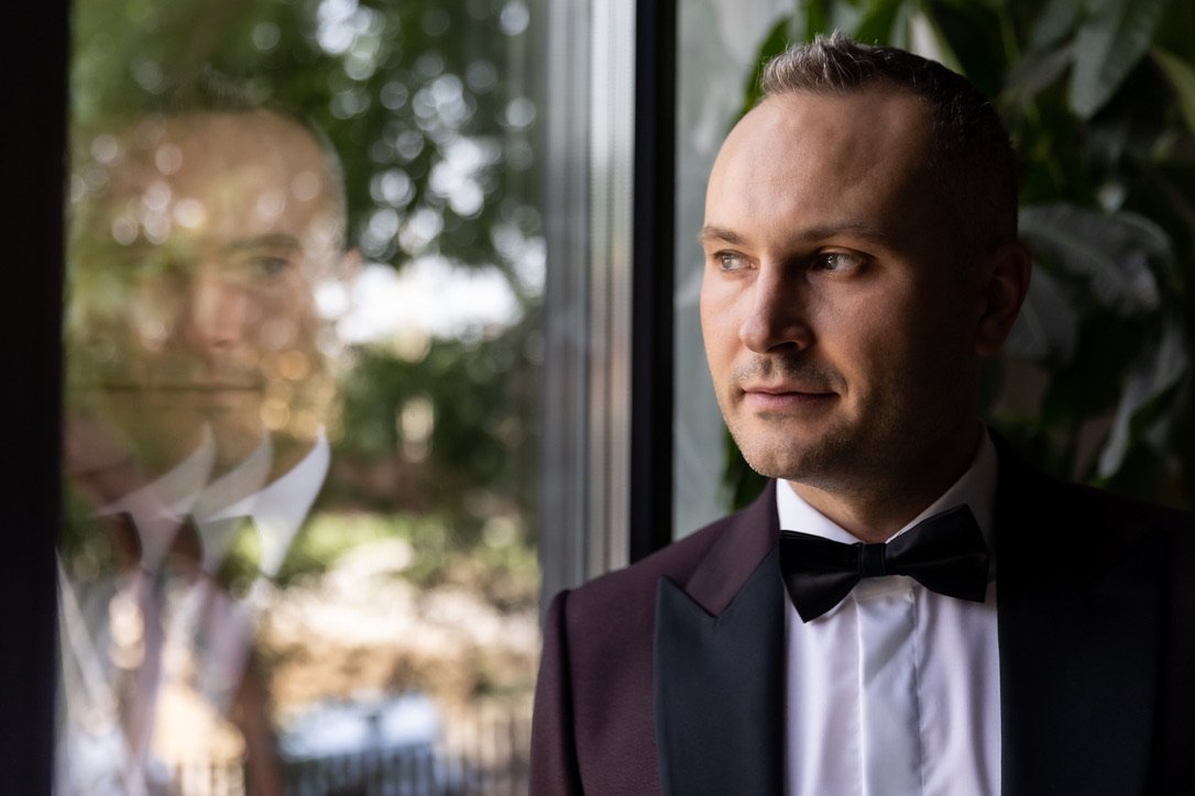 A man in a tuxedo with a bow tie looking out a window with a reflection of himself visible on the glass.