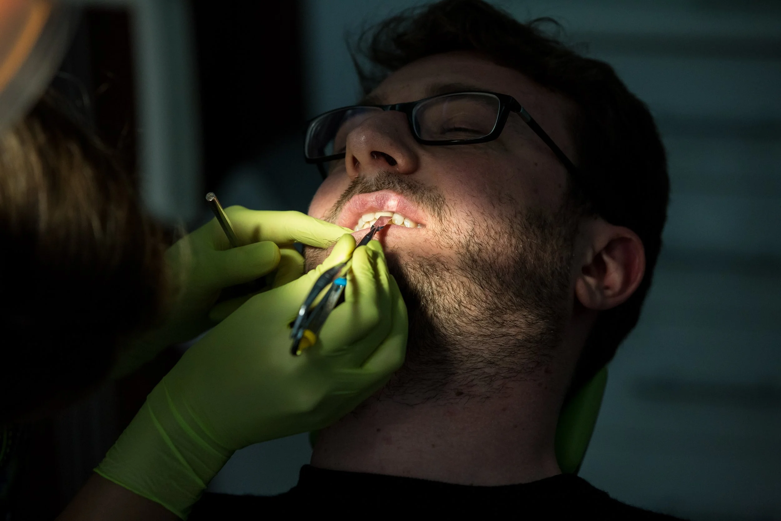 A person with dark glasses and facial hair receives dental treatment from a dentist wearing green gloves, using dental tools in a dark room.