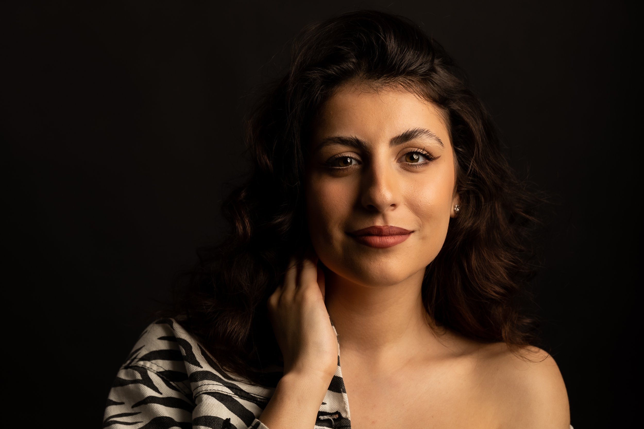 A young woman with wavy dark hair and brown eyes, looking at the camera with a slight smile, wearing a zebra-patterned top and earrings, against a black background.