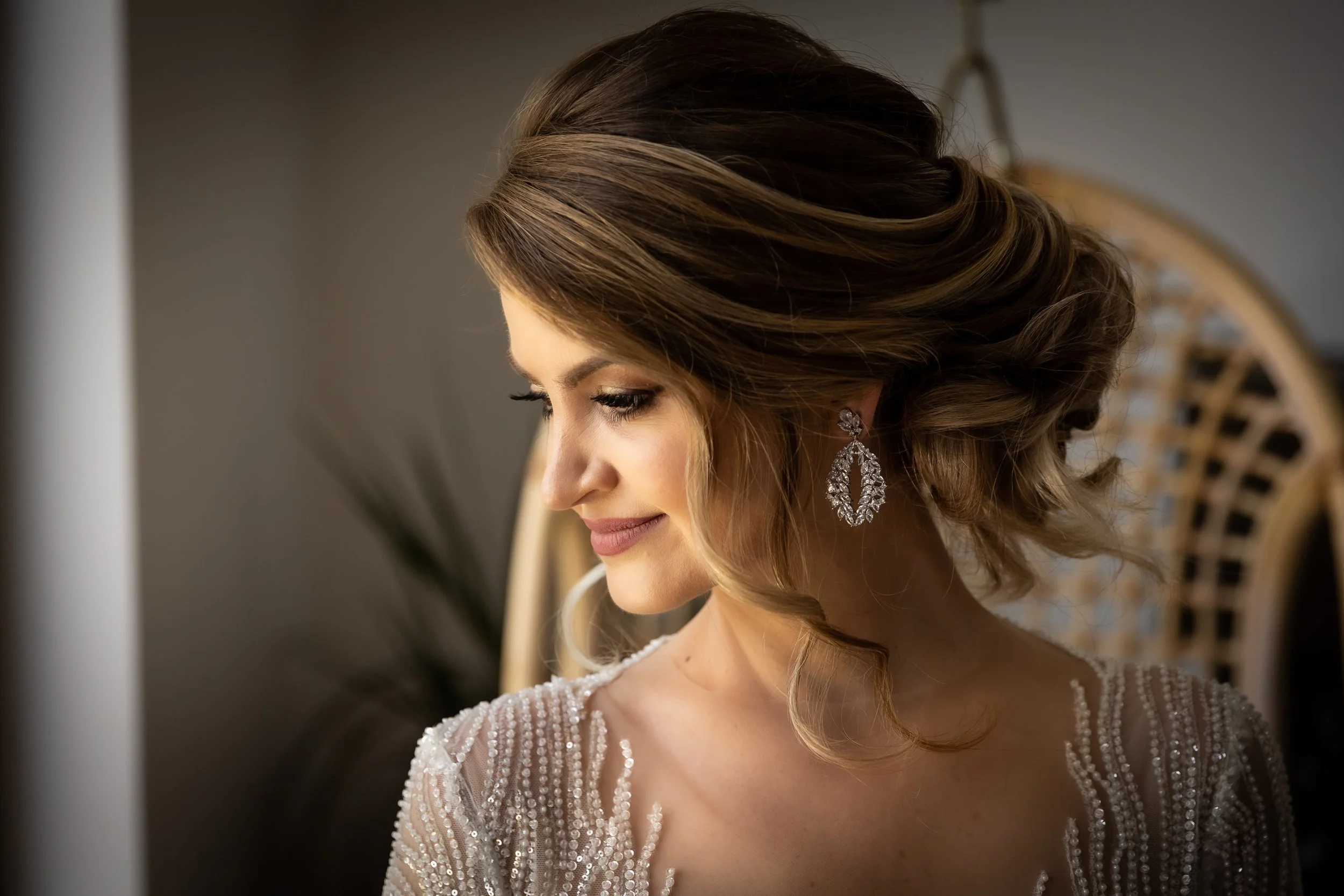 Close-up of a bride with styled brown hair, wearing chandelier earrings and a beaded dress, looking down, softly smiling.