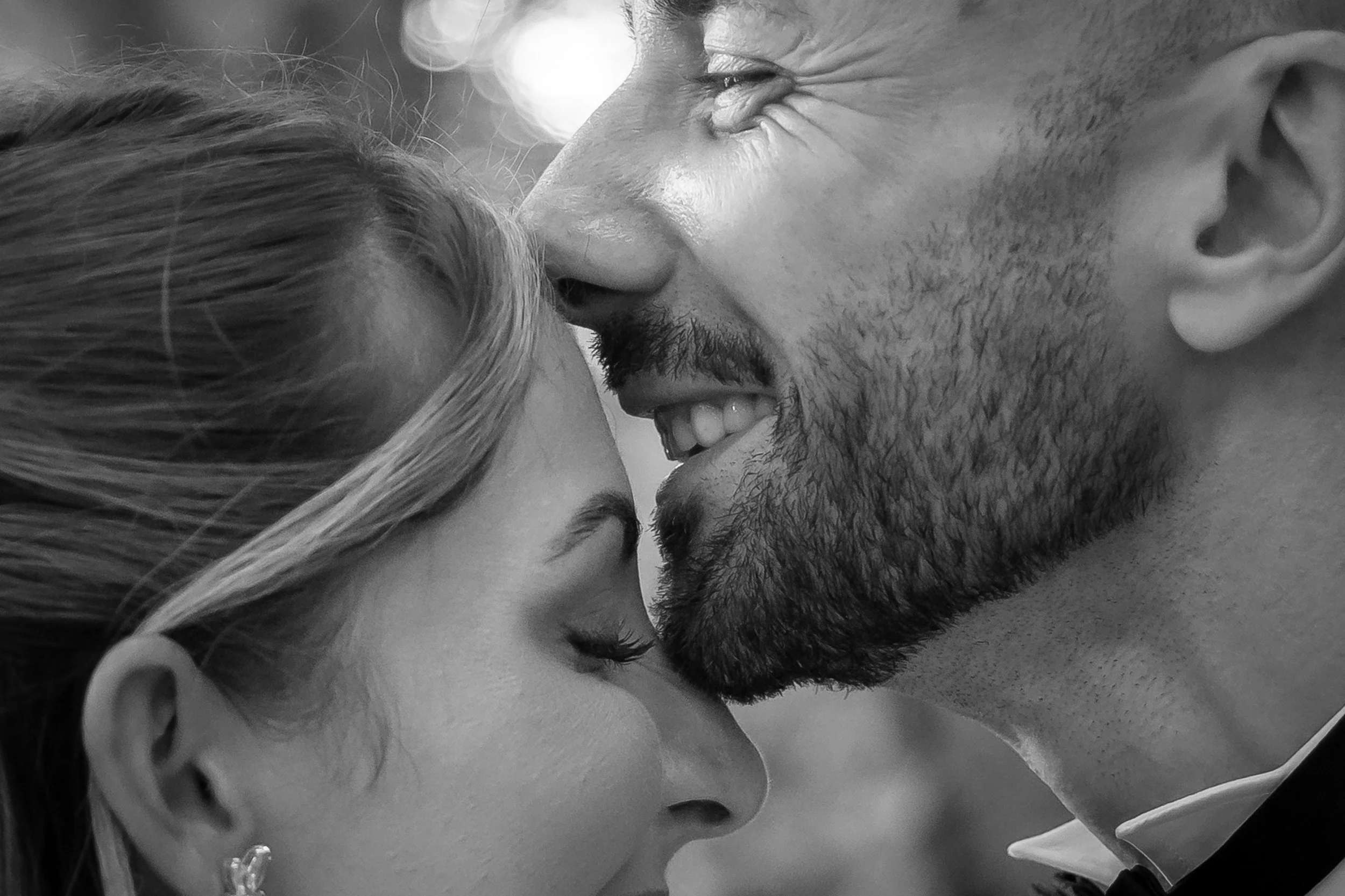A close-up black and white photo of a man and woman sharing an intimate moment, with their faces close together, eyes closed, smiling, and forehead touching.