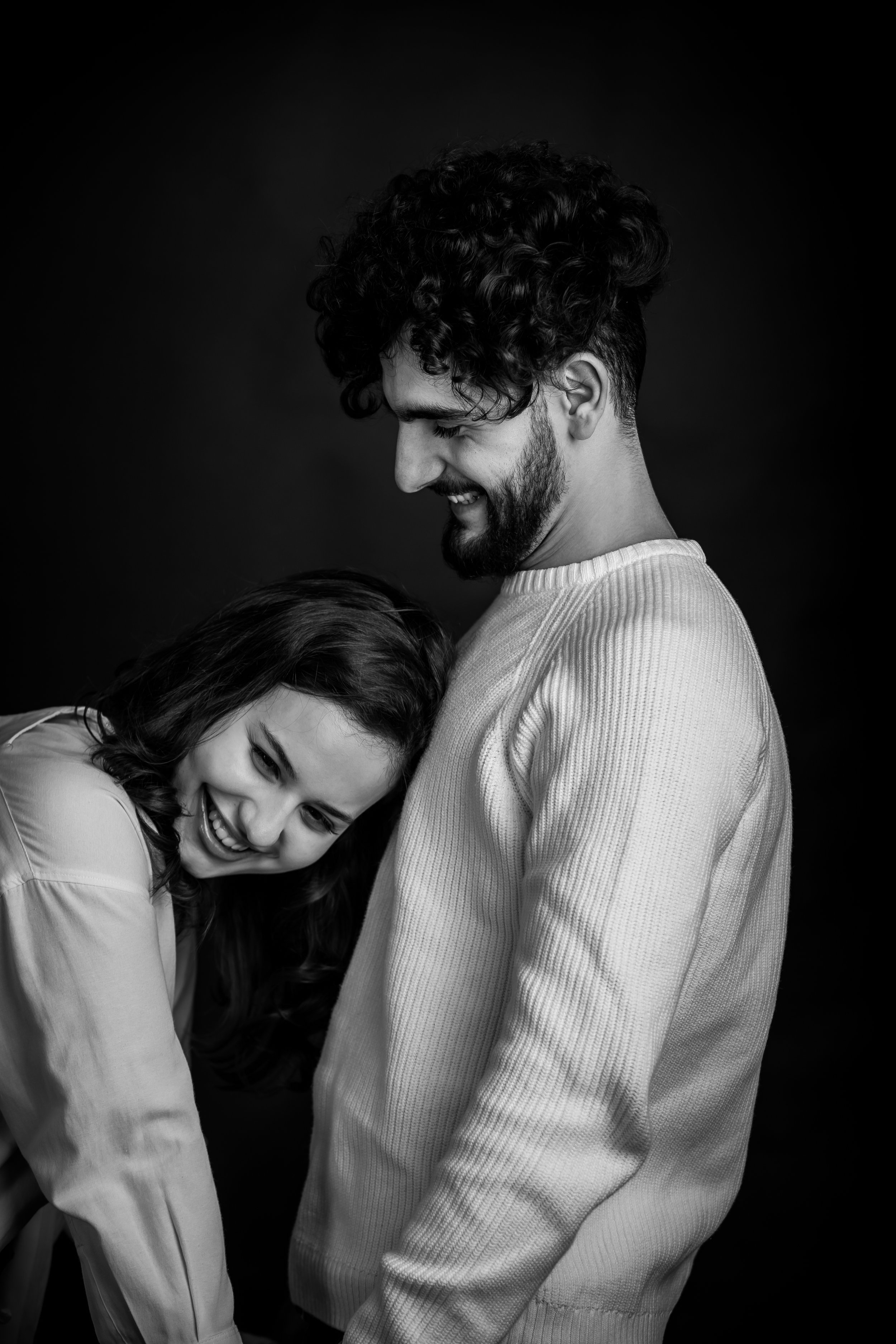 A black-and-white photo of a smiling man with curly hair and a beard, standing close to a woman with long hair who is leaning her head on his chest, both appearing happy and affectionate.