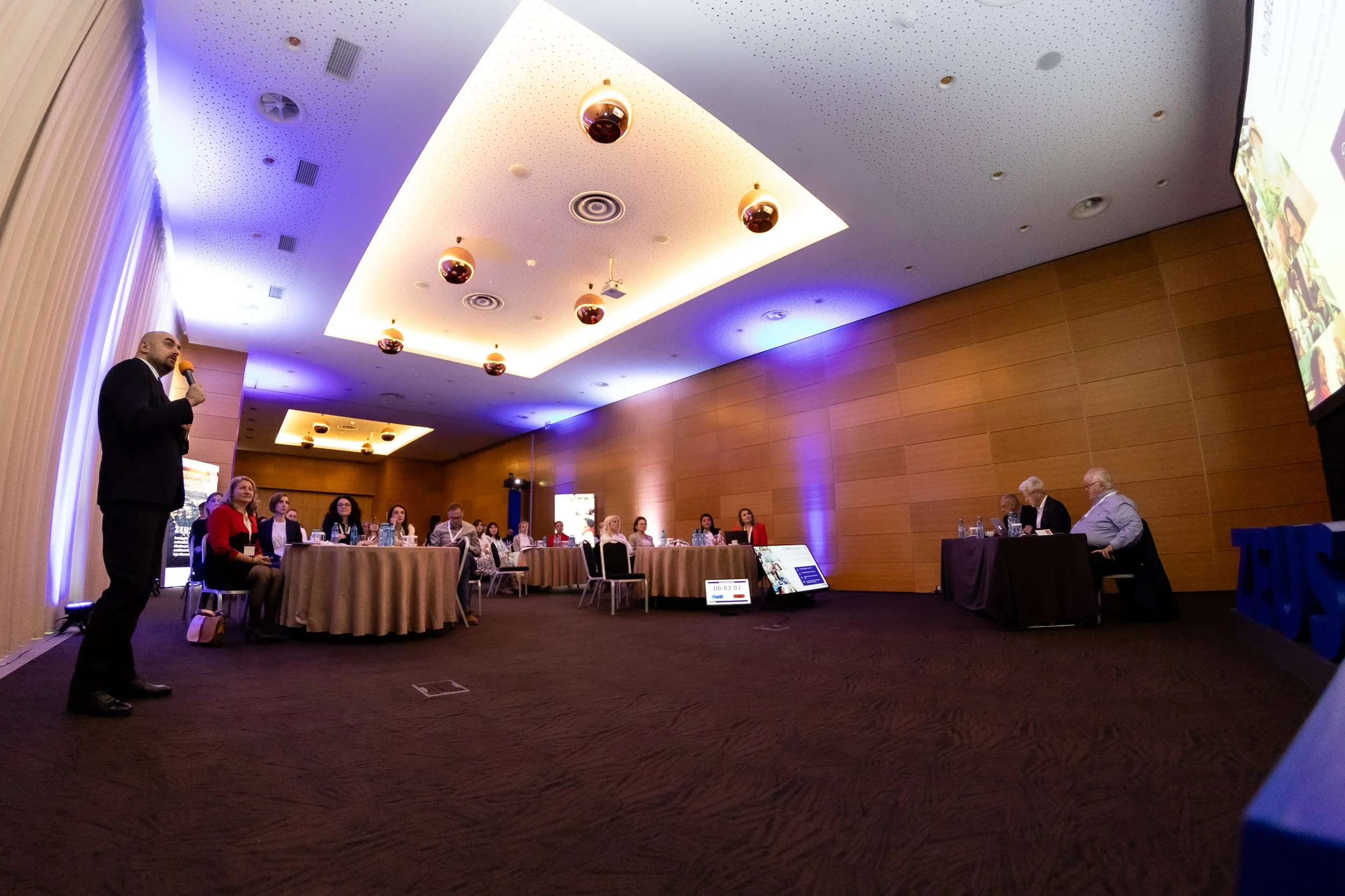 A conference room with a man speaking into a microphone on the left side and a panel of people seated at a table on the right side. The room has wooden walls and a ceiling with embedded lights and speakers. There is a large screen on the right side d