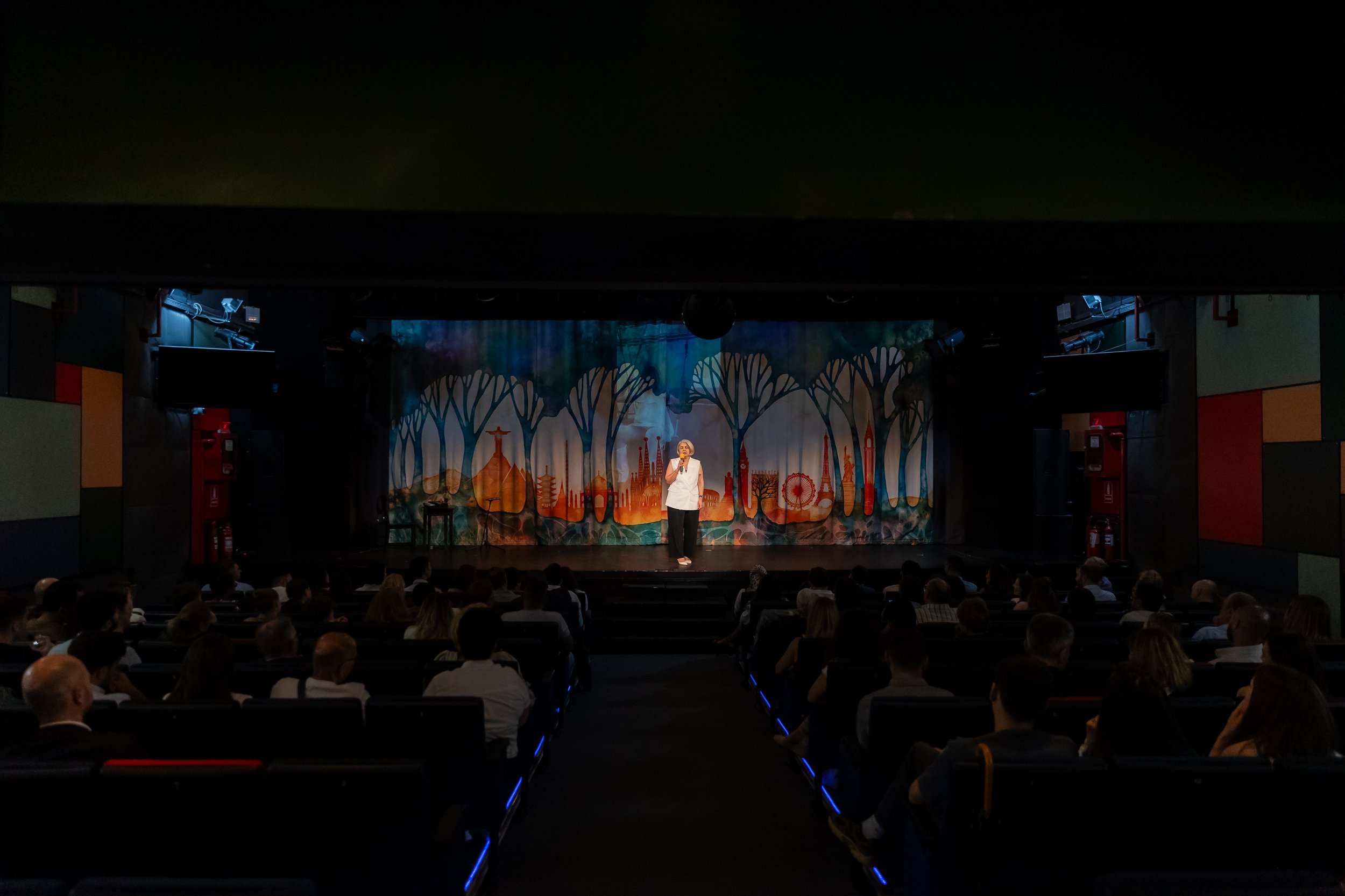 A woman is standing on stage in a theater or performance venue, speaking to an audience. The stage has a colorful backdrop with abstract tree and cityscape designs, and there are multiple audience members seated and listening.