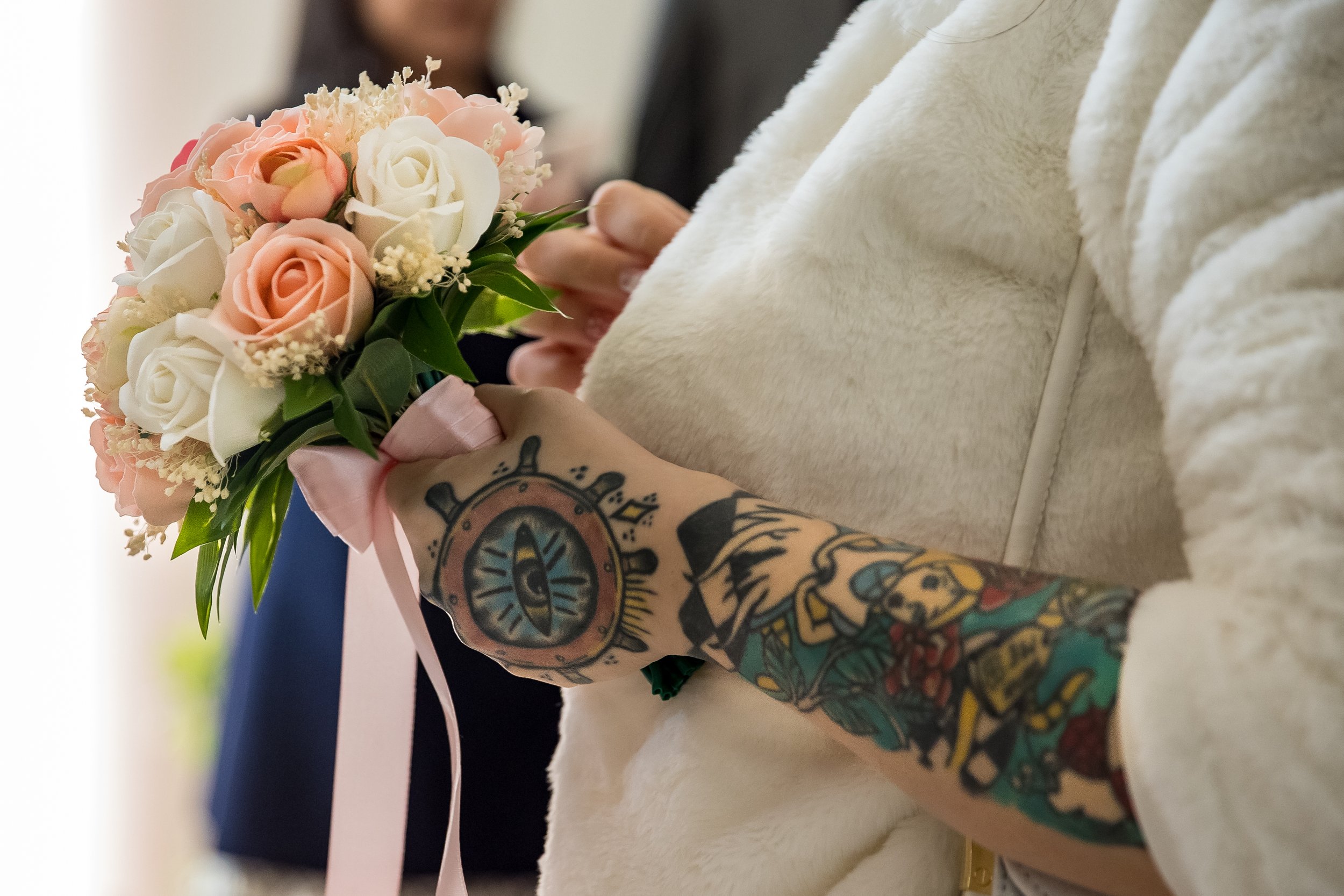 A person with tattooed arms holding a bouquet of pink and white roses. The person is wearing a light-colored, soft-looking garment and is sitting on a white surface.