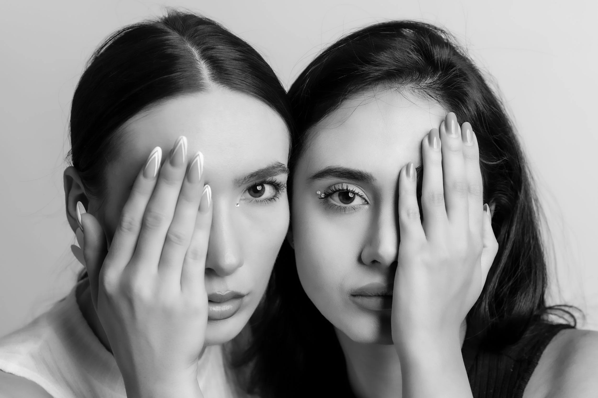 Two women with dark hair and light skin, covering one eye with their hands, looking directly at the camera in black and white.