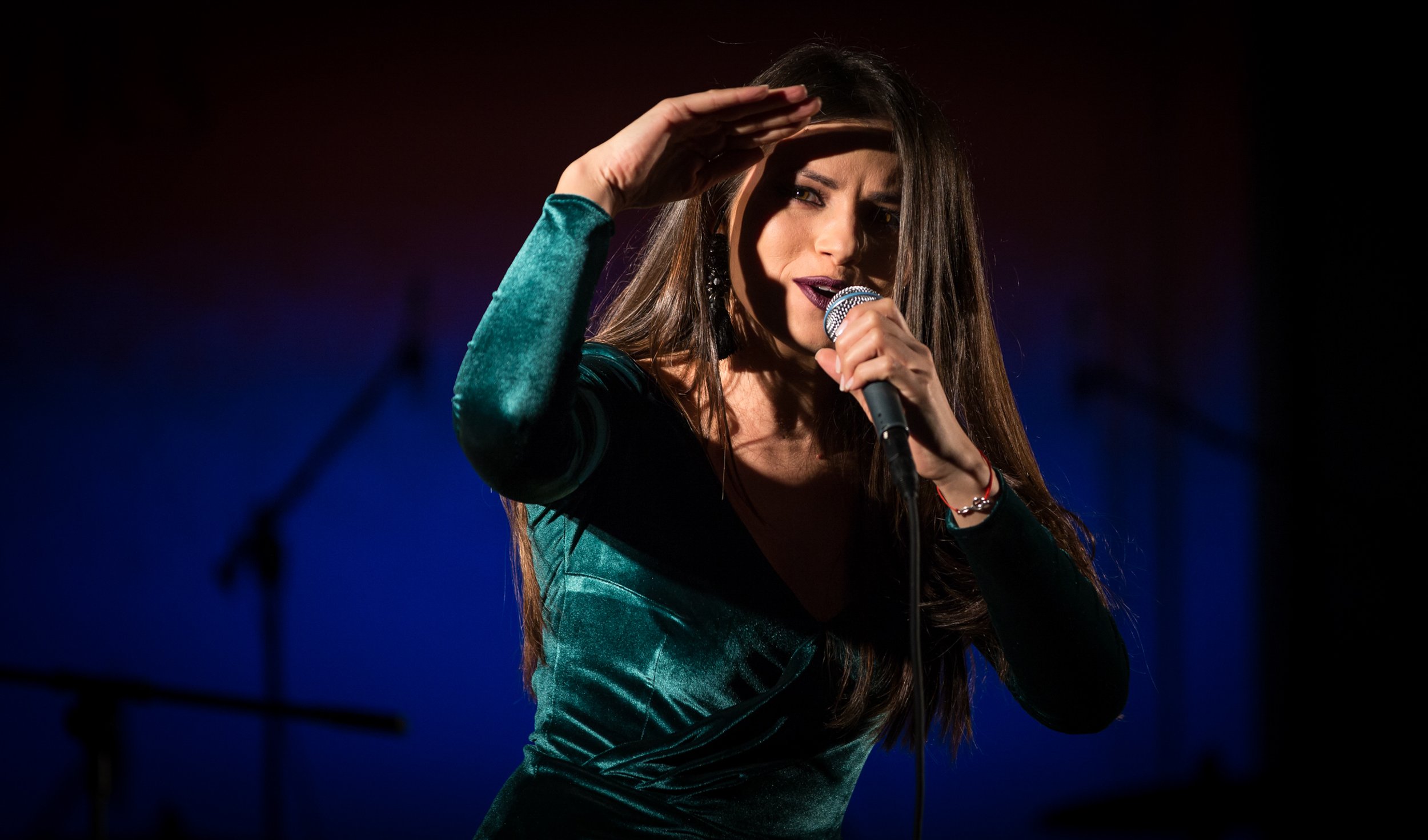 A woman with long brown hair wearing a teal dress with dark green velvet fabric, holding a microphone in her right hand, and shielding her eyes with her left hand against stage lights.