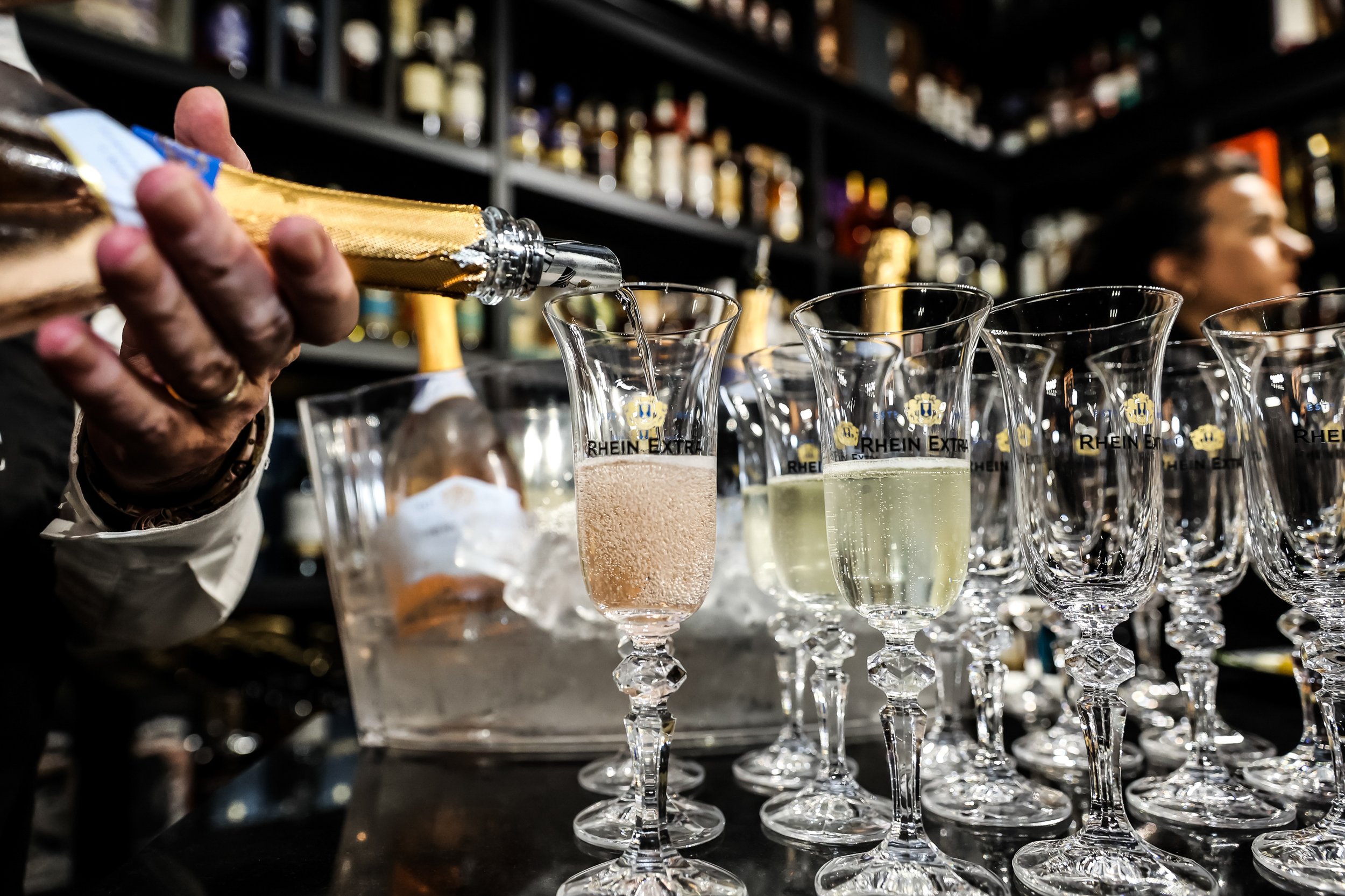 A bartender pours champagne into flutes on a bar with shelves of liquor bottles in the background.