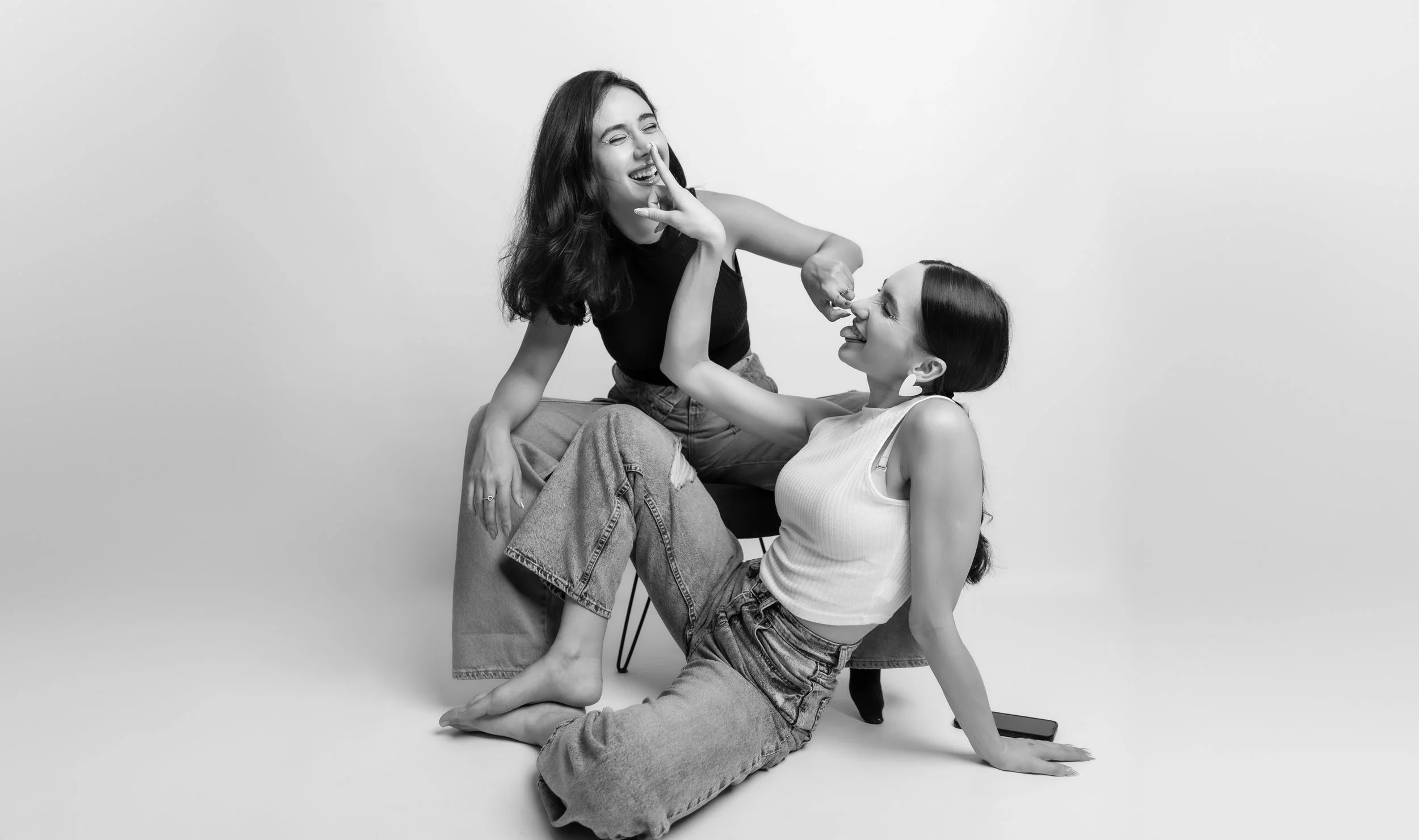 Two young women with dark hair, one sitting on a chair and the other on the floor, engaging in playful gesture and smiling, in black and white photography.