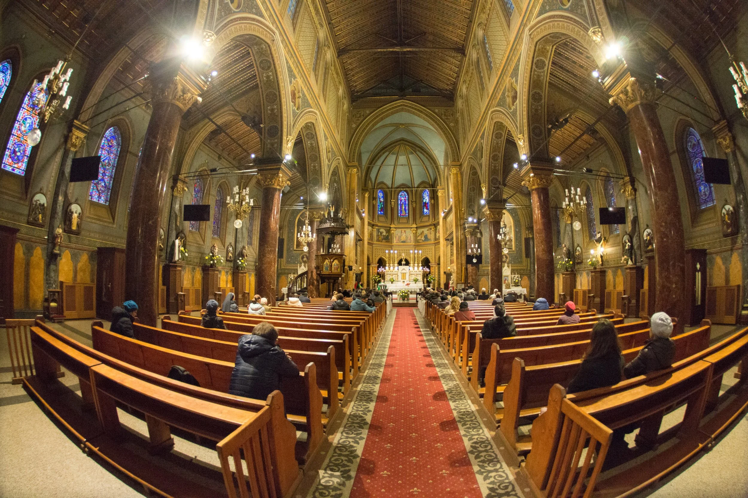 Interior of a church with rows of wooden pews, stained glass windows, marble columns, and an altar at the front, filled with people seated and attending service.
