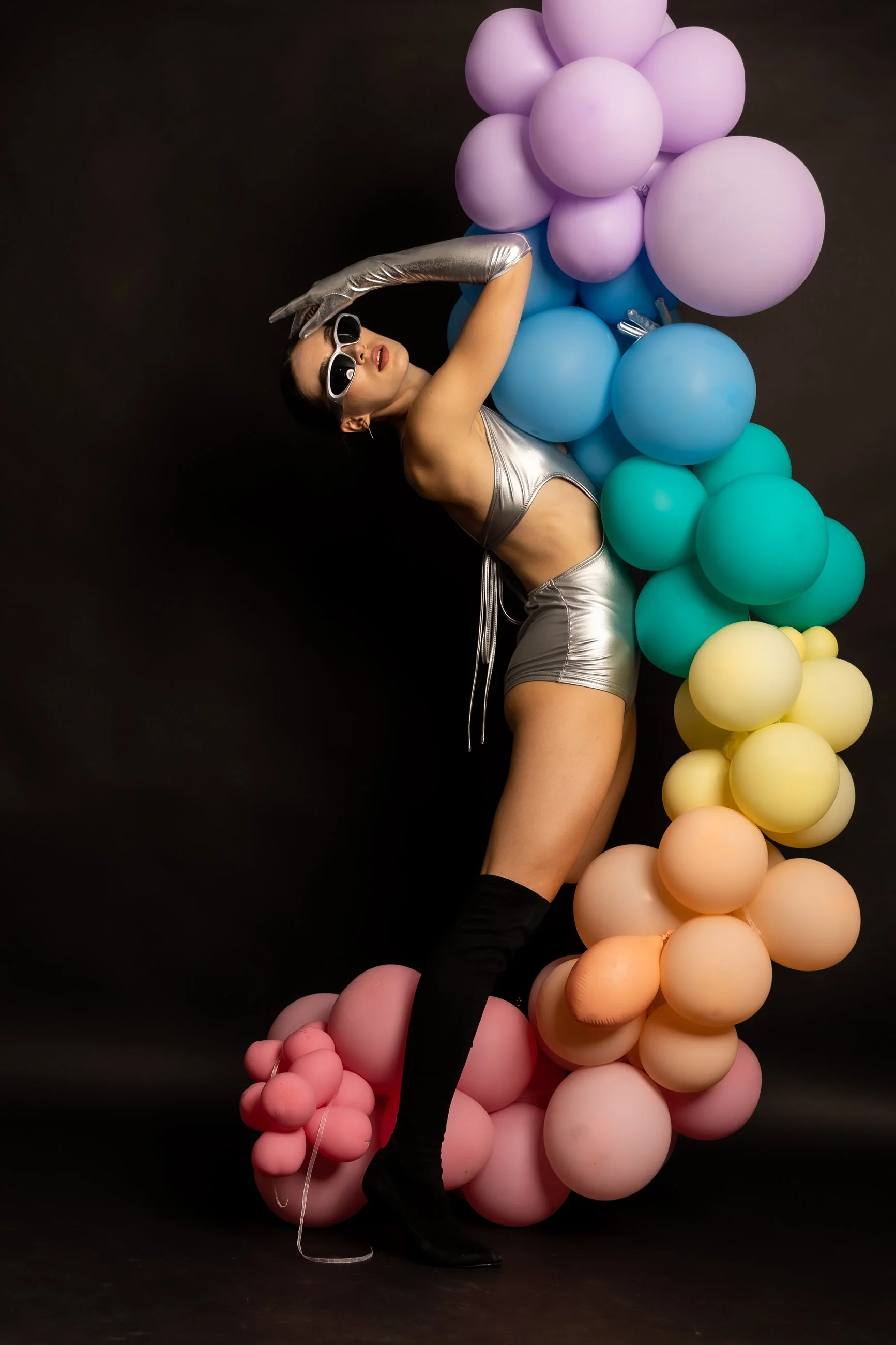 A woman in a silver metallic outfit with white sunglasses poses against a dark background. She is surrounded by colorful balloons arranged in a cascading pattern, including purple, blue, green, yellow, orange, pink, and peach, creating a vibrant and 
