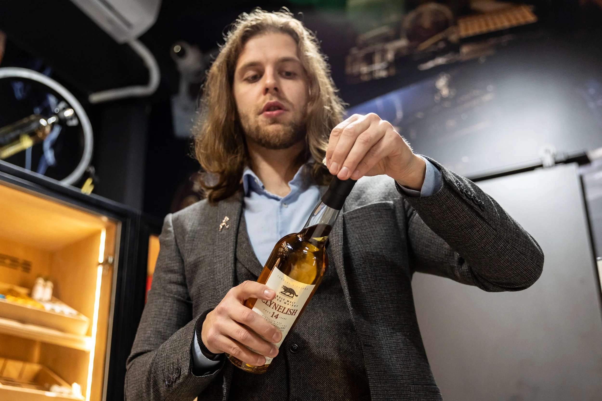 A man with long hair and a beard opening a bottle of whiskey in a dimly lit room, with a wine fridge in the background.