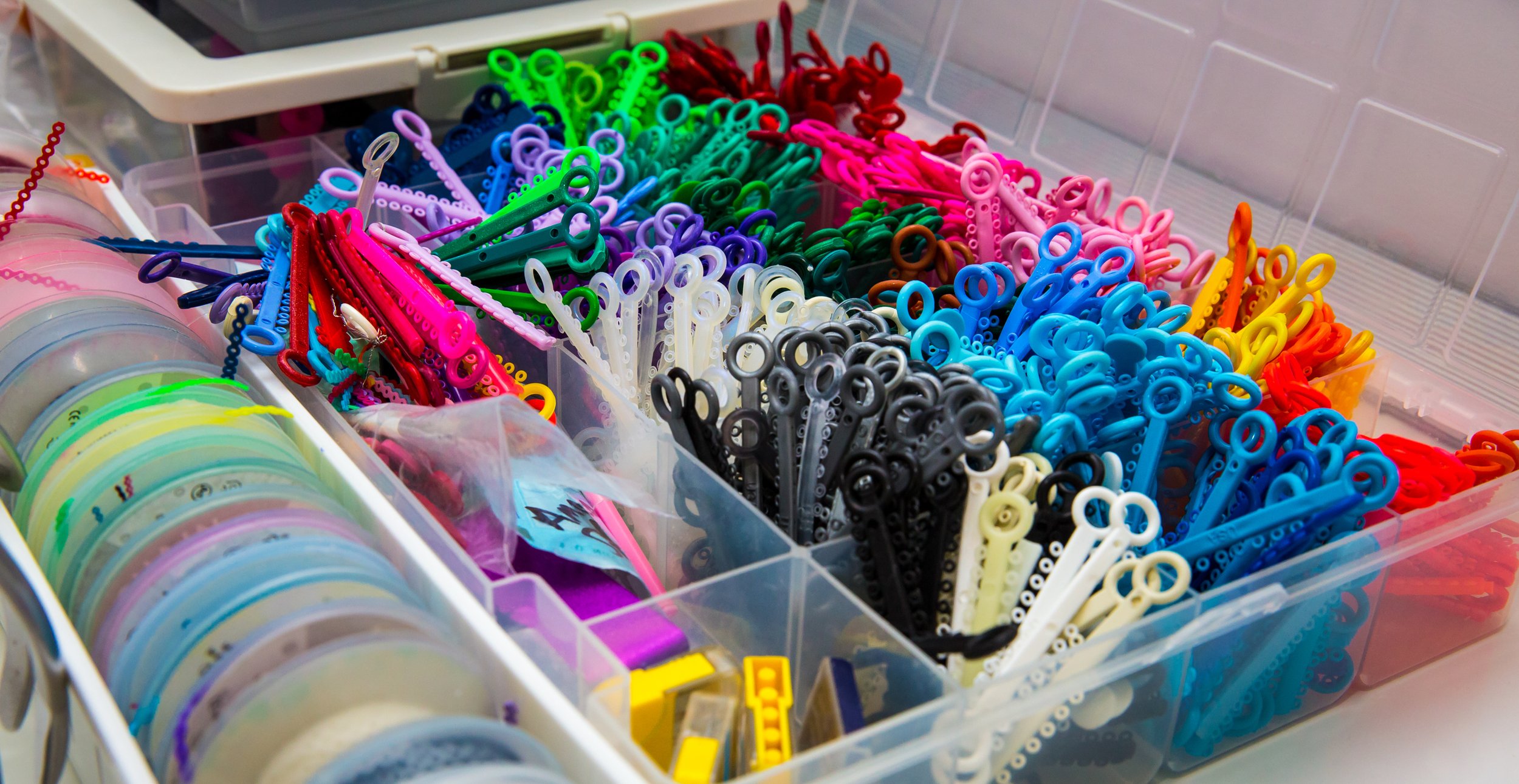 A plastic storage box filled with colorful hair ties and hair clips in various colors, including red, pink, purple, green, blue, black, white, yellow, and orange.