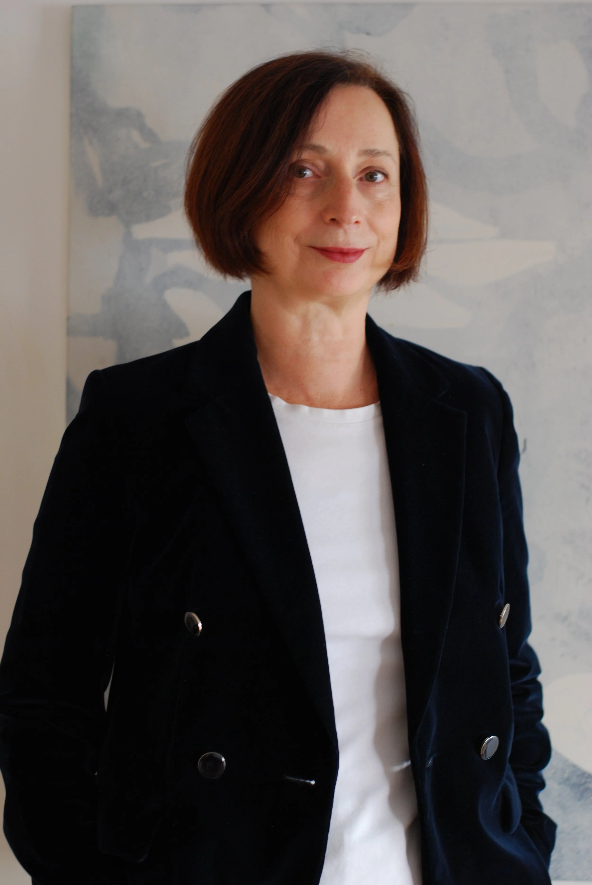 A woman with short brown hair, wearing a black blazer, white shirt, and standing against an abstract blue and white background.