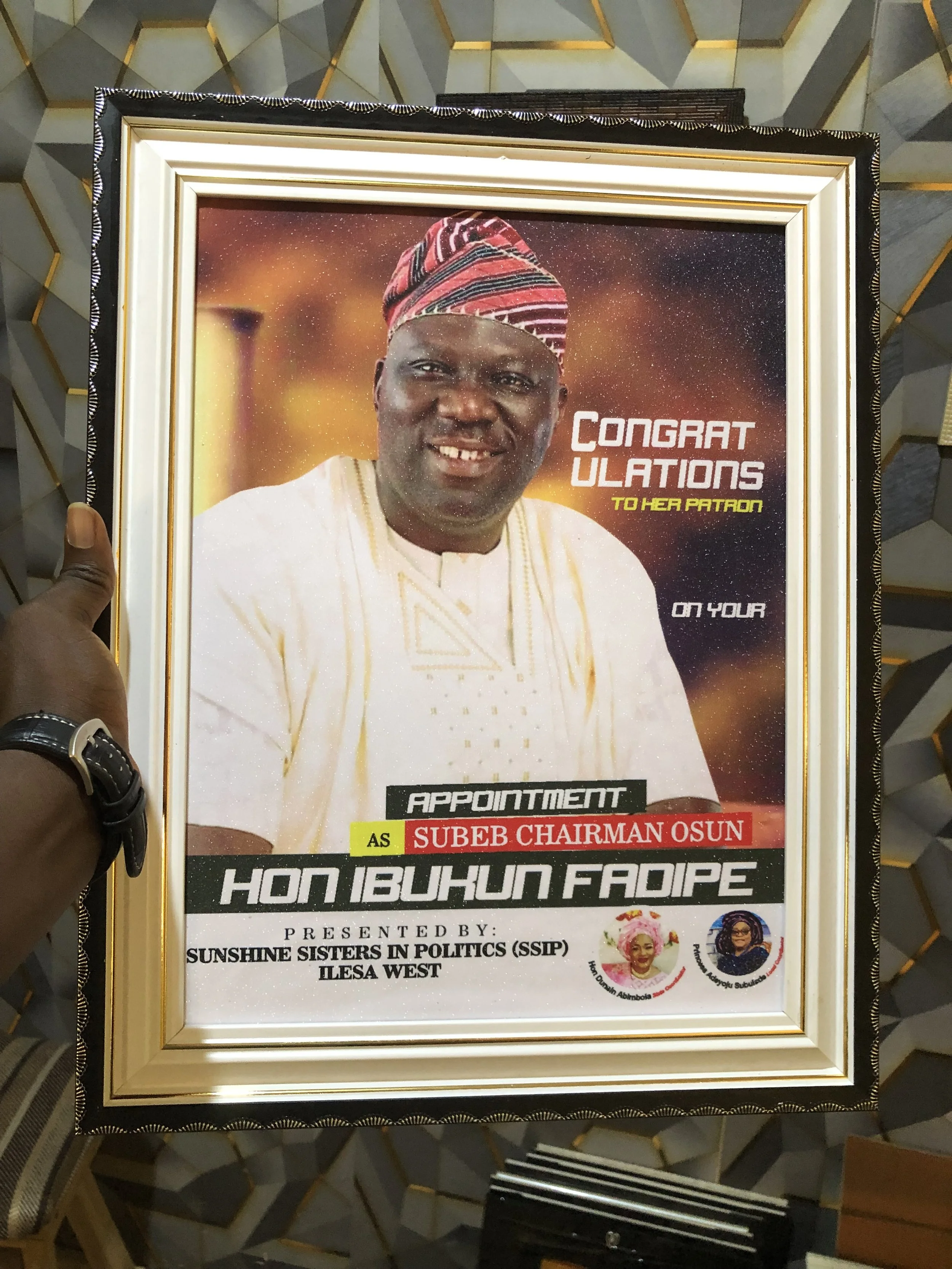 A framed congratulatory poster featuring a man wearing traditional West African attire and a cap, with text indicating an appointment to a political position, presented by a women's political group.