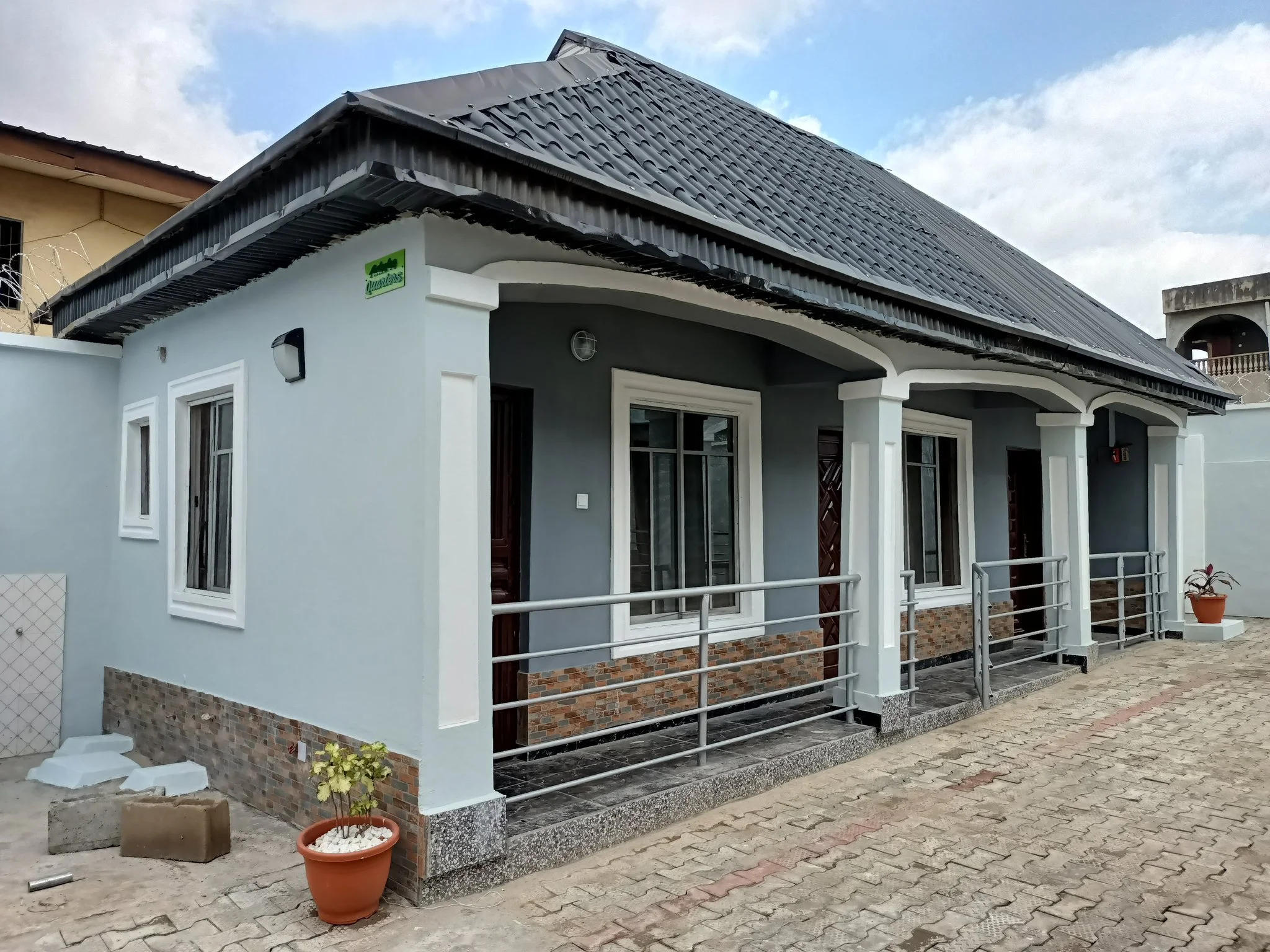 A modern house with a white exterior, large windows, and a steel roof. There is a small front porch with a railing and two potted plants in front. The house is situated on a paved area with some construction materials nearby.