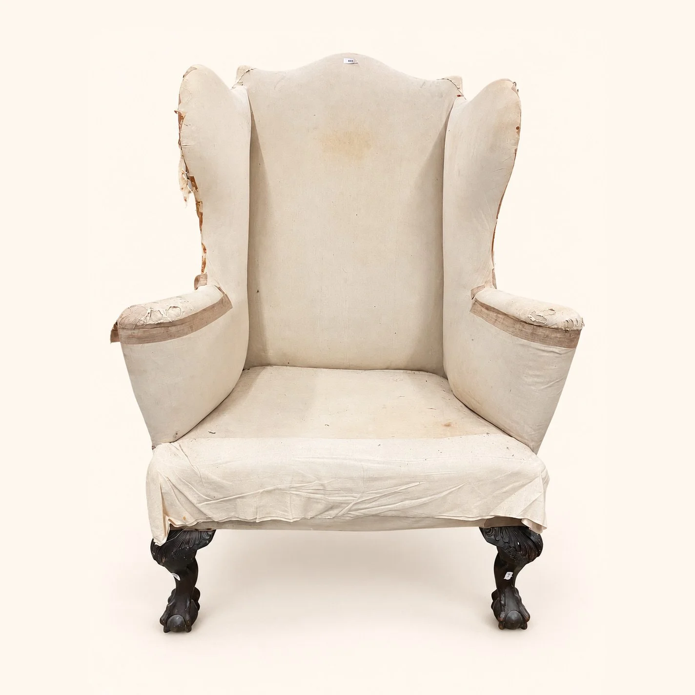Before reupholstery: a tall wingback chair in worn cream fabric, with curved arms and dark carved wooden legs.