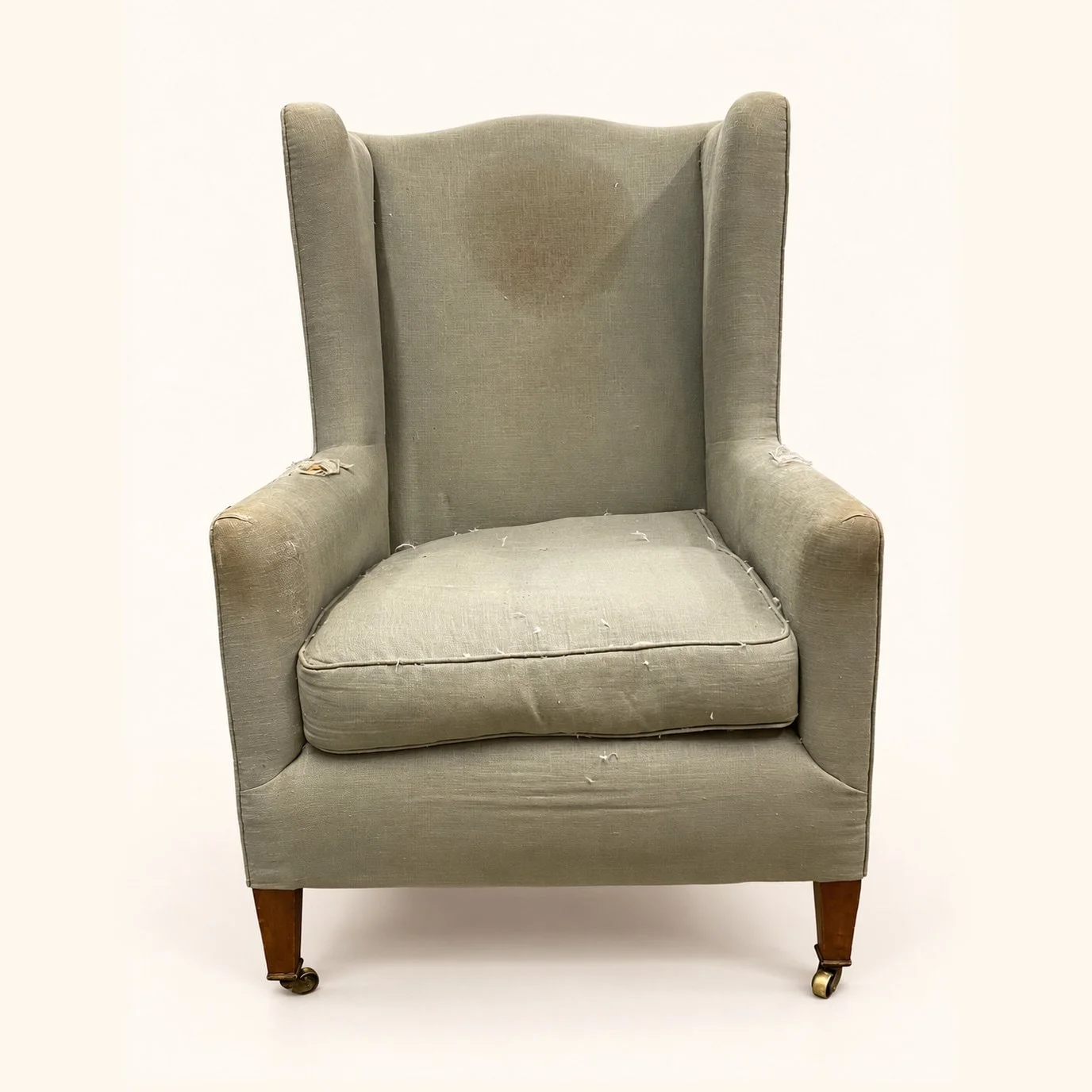 William Birch Wingback Chair before reupholstery in worn plain grey-green fabric, with wooden legs and front castors.