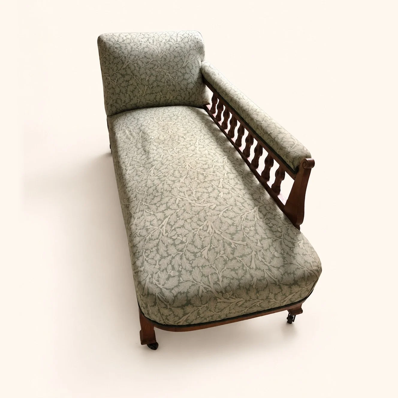 Chaise longue before reupholstery, with sage leaf-patterned fabric, a wooden spindle side and castor feet.