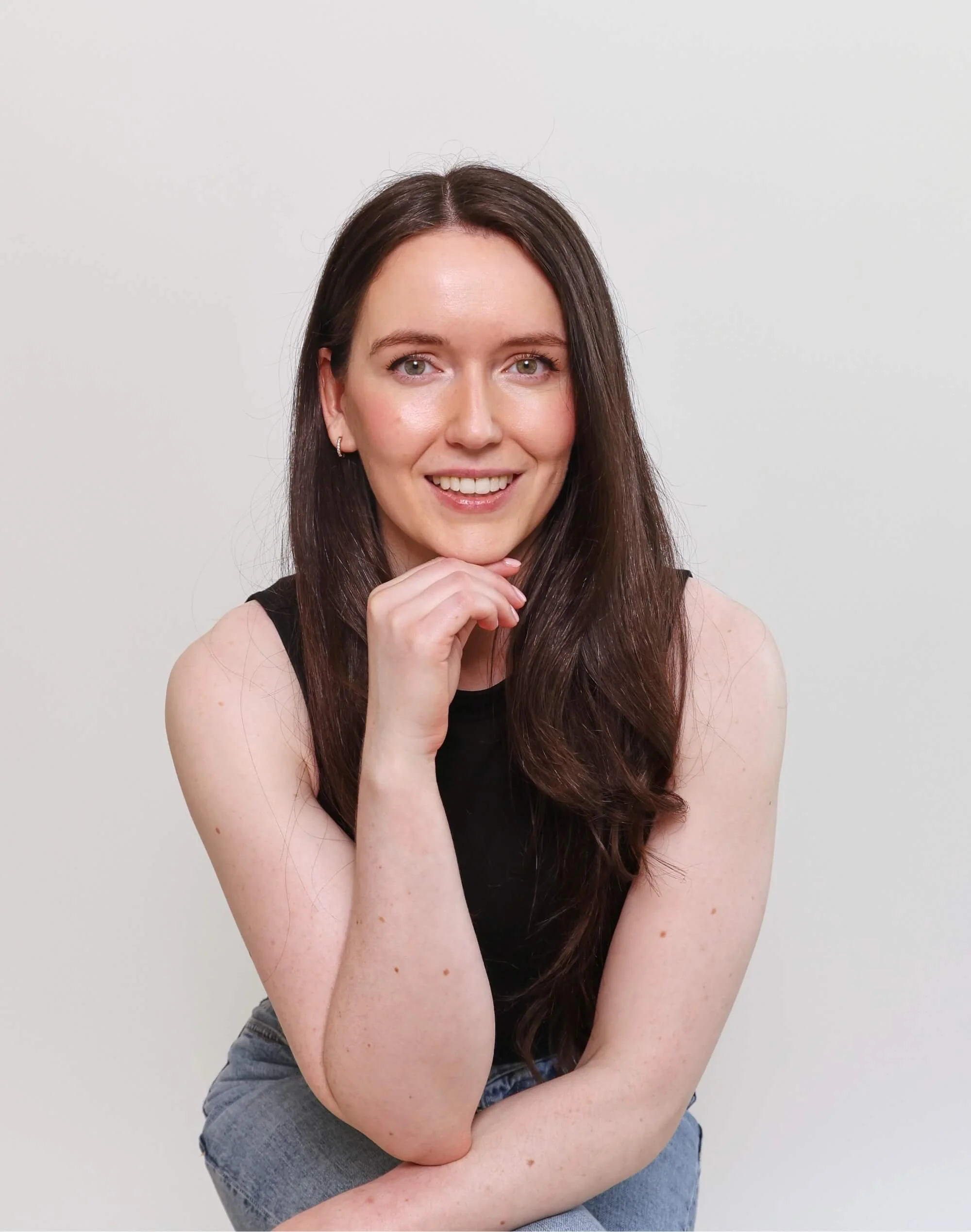 A young woman with long brown hair, green eyes, and fair skin, smiling, wearing a sleeveless black top and blue jeans, posing against a plain white background.