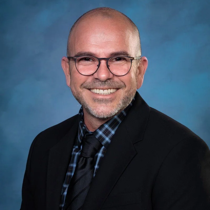 Professional headshot of Alex Hamby smiling, wearing black-framed glasses, a black suit and tie, and a blue plaid shirt against a blue background.