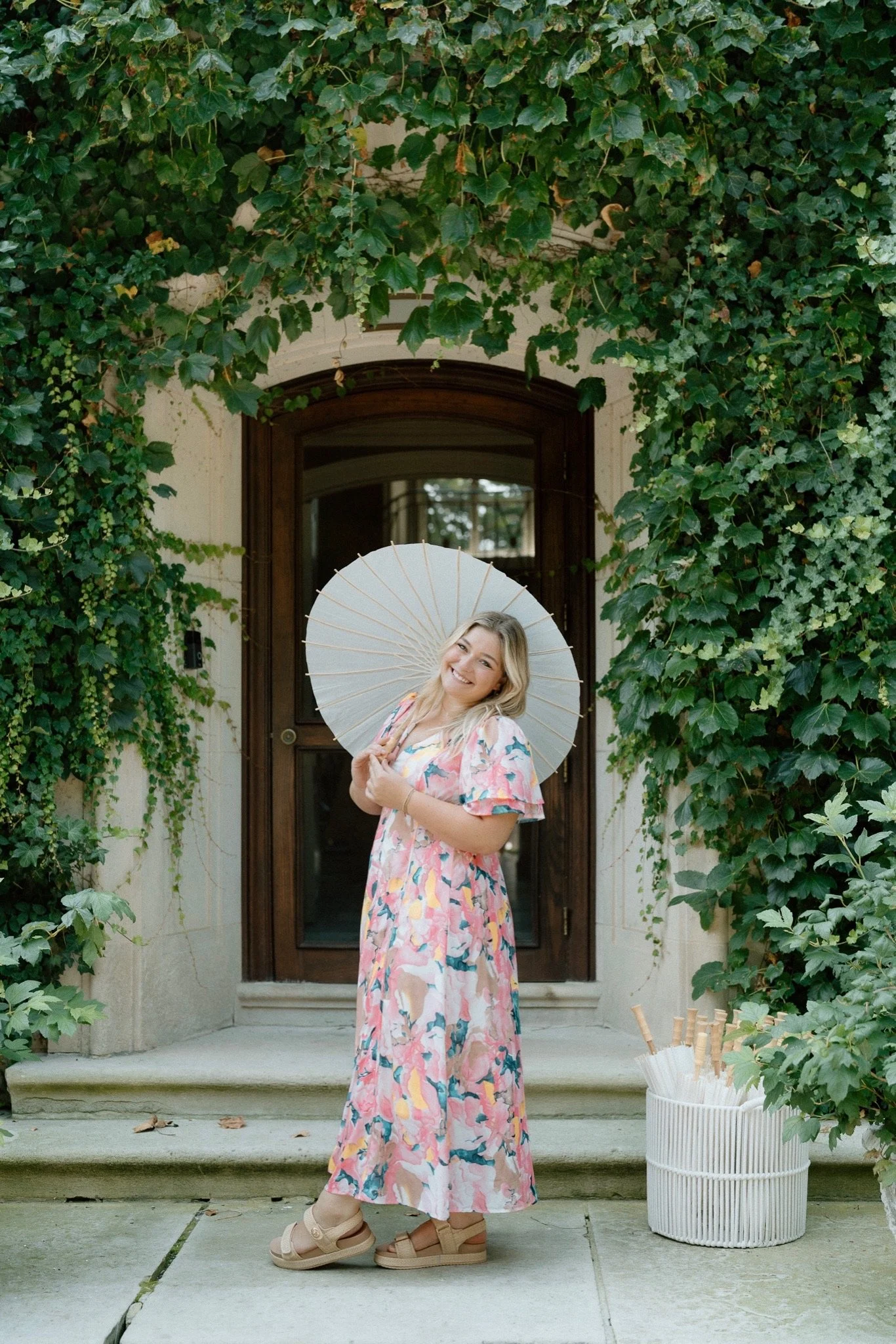 A young woman with blonde hair holding a white parasol, smiling, in front of a wooden door surrounded by greenery, wearing a floral dress and beige platform sandals, with a white basket of rolled-up paper umbrellas nearby.