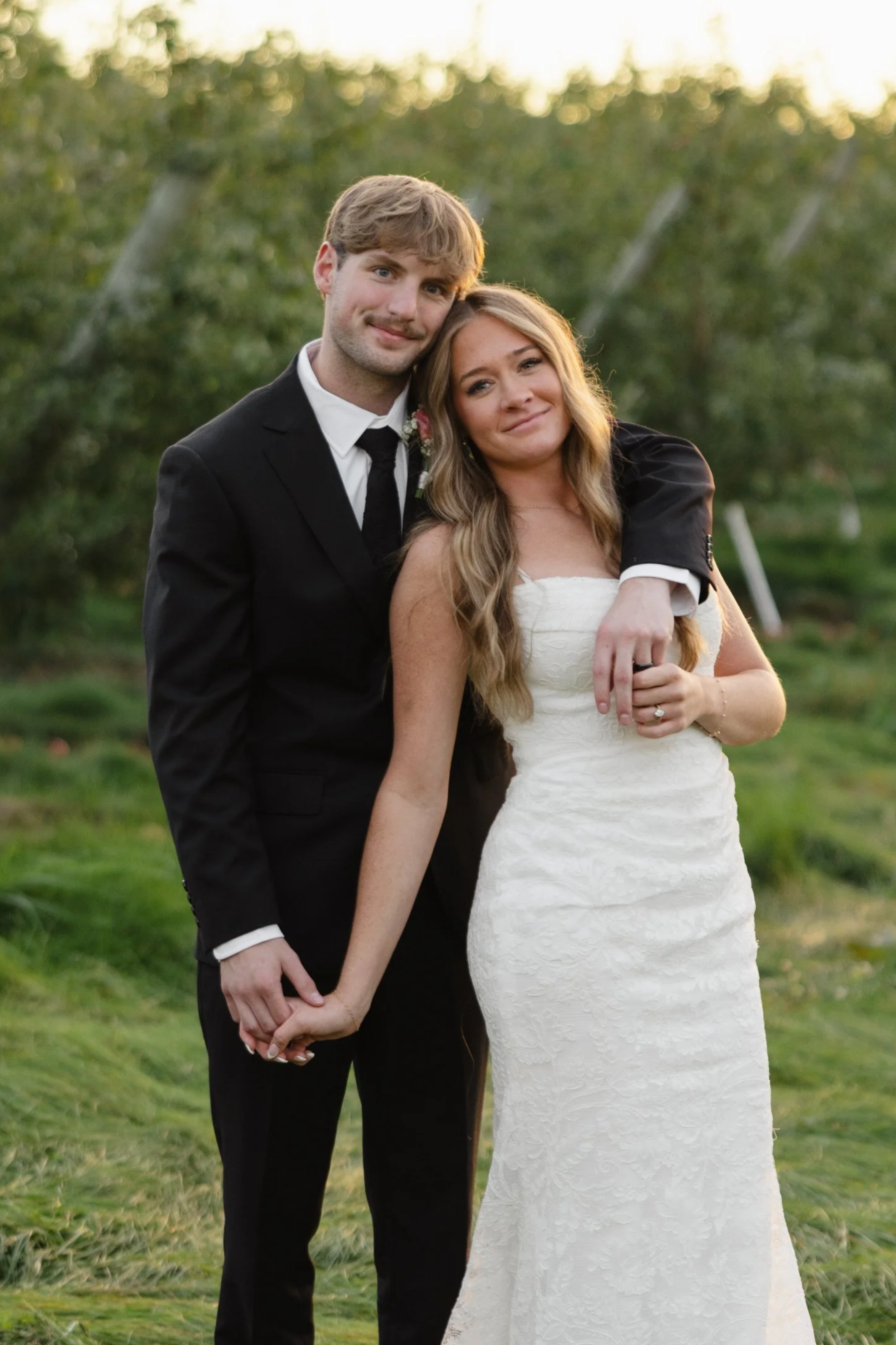 A newlywed couple stands together outside on a grassy area with trees in the background during sunset. The groom, dressed in a black suit and tie, has his arm around the bride's shoulders. The bride is wearing a strapless white wedding gown and has her hand resting on the groom's hand, showing her wedding ring. They are smiling and holding hands, looking happy and content.