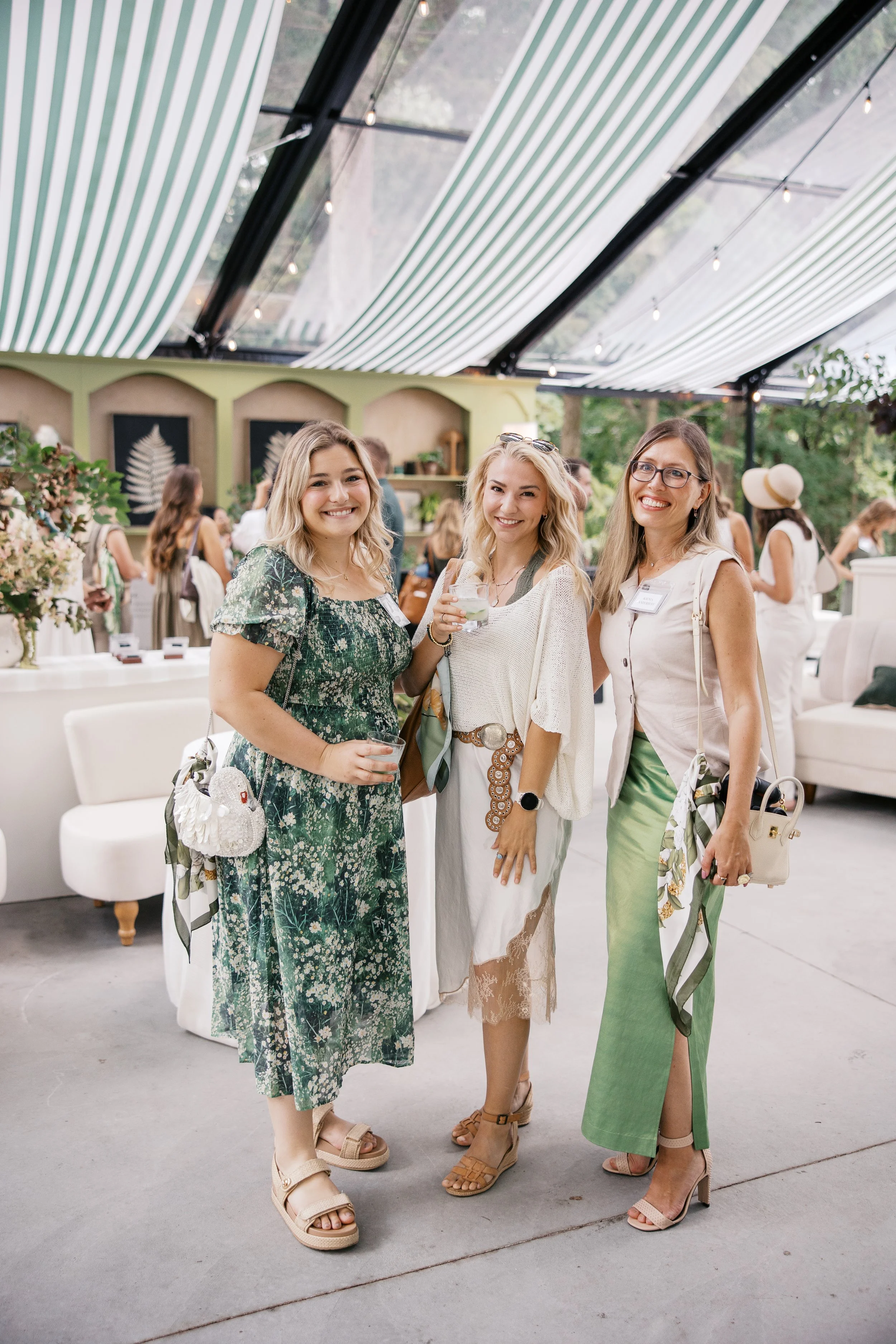 Three smiling women wearing summer dresses and sandals standing together at an indoor event, with other guests and decorative plants in the background.