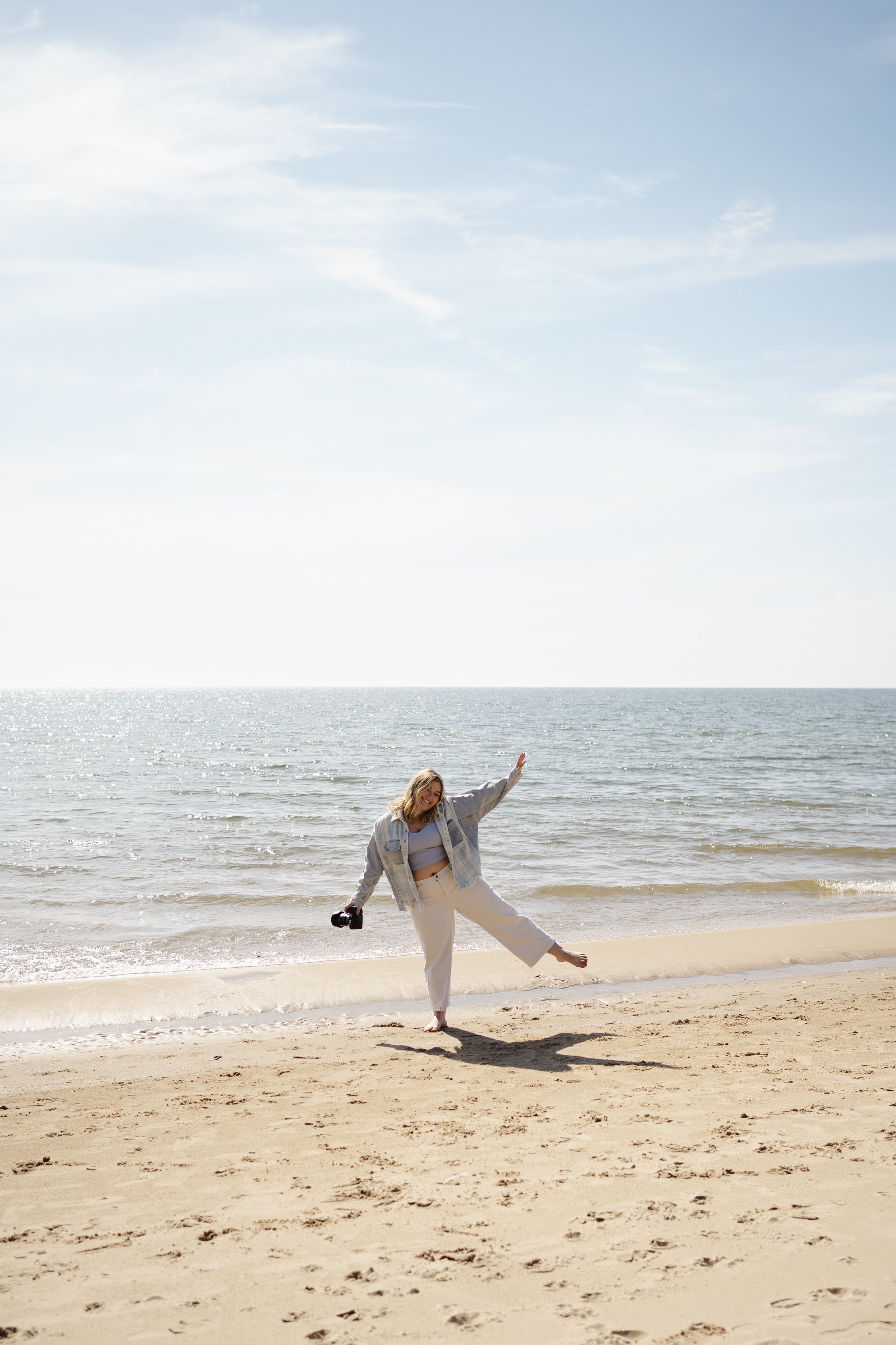 A woman standing on a sandy beach holding a camera and posing, with the ocean and sky in the background, and others in the distance.