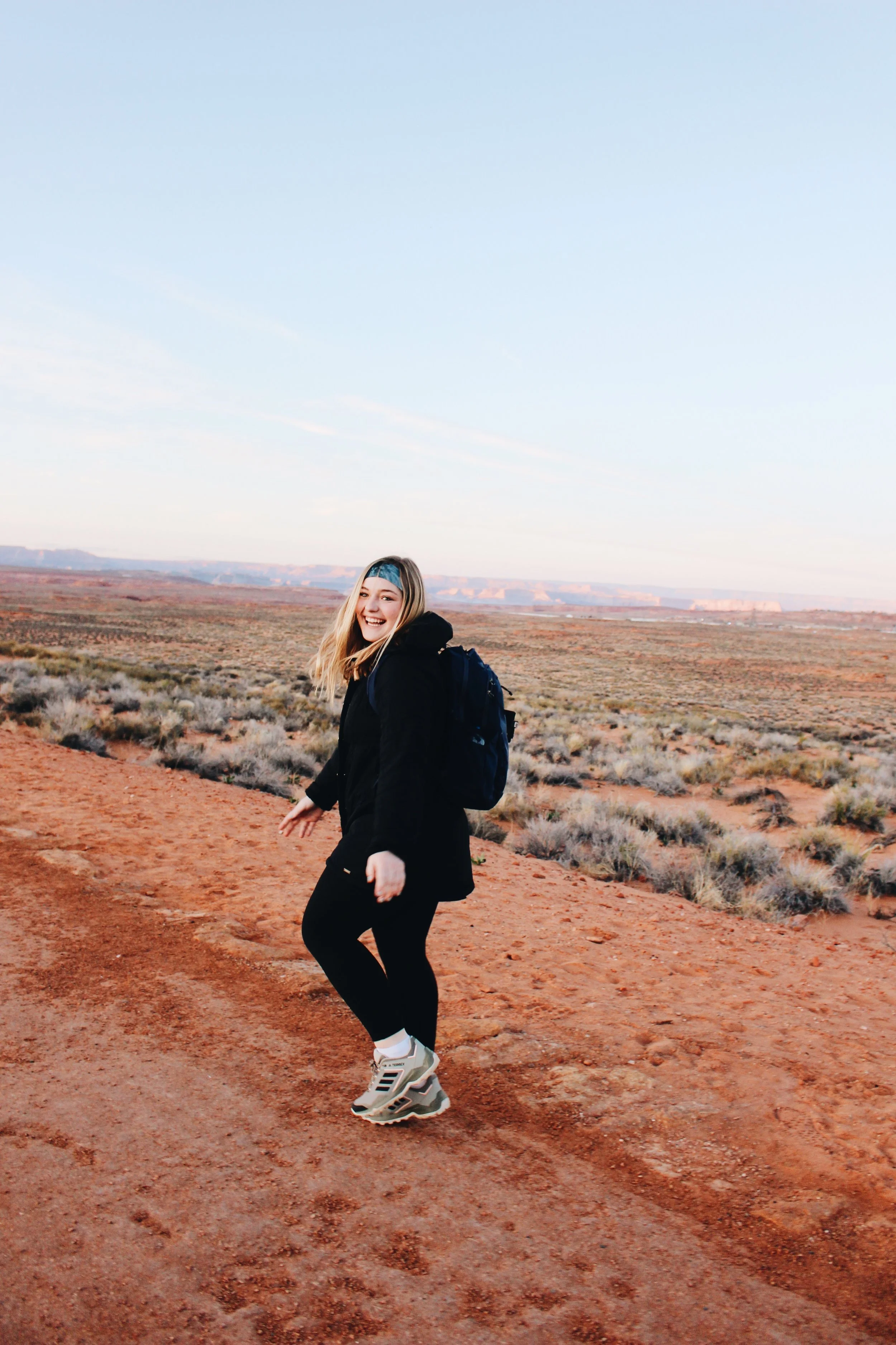 A woman with long blonde hair, wearing a black jacket, black pants, and gray sneakers, smiling and looking back while walking on a dirt trail in a desert landscape with sparse vegetation and distant red canyon formations.