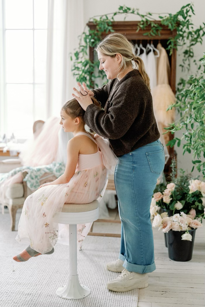 A woman in a dark sweater and jeans styles a young girl in a light pink dress with a tulle skirt in a bright, flower-decorated room, possibly preparing for a special occasion.