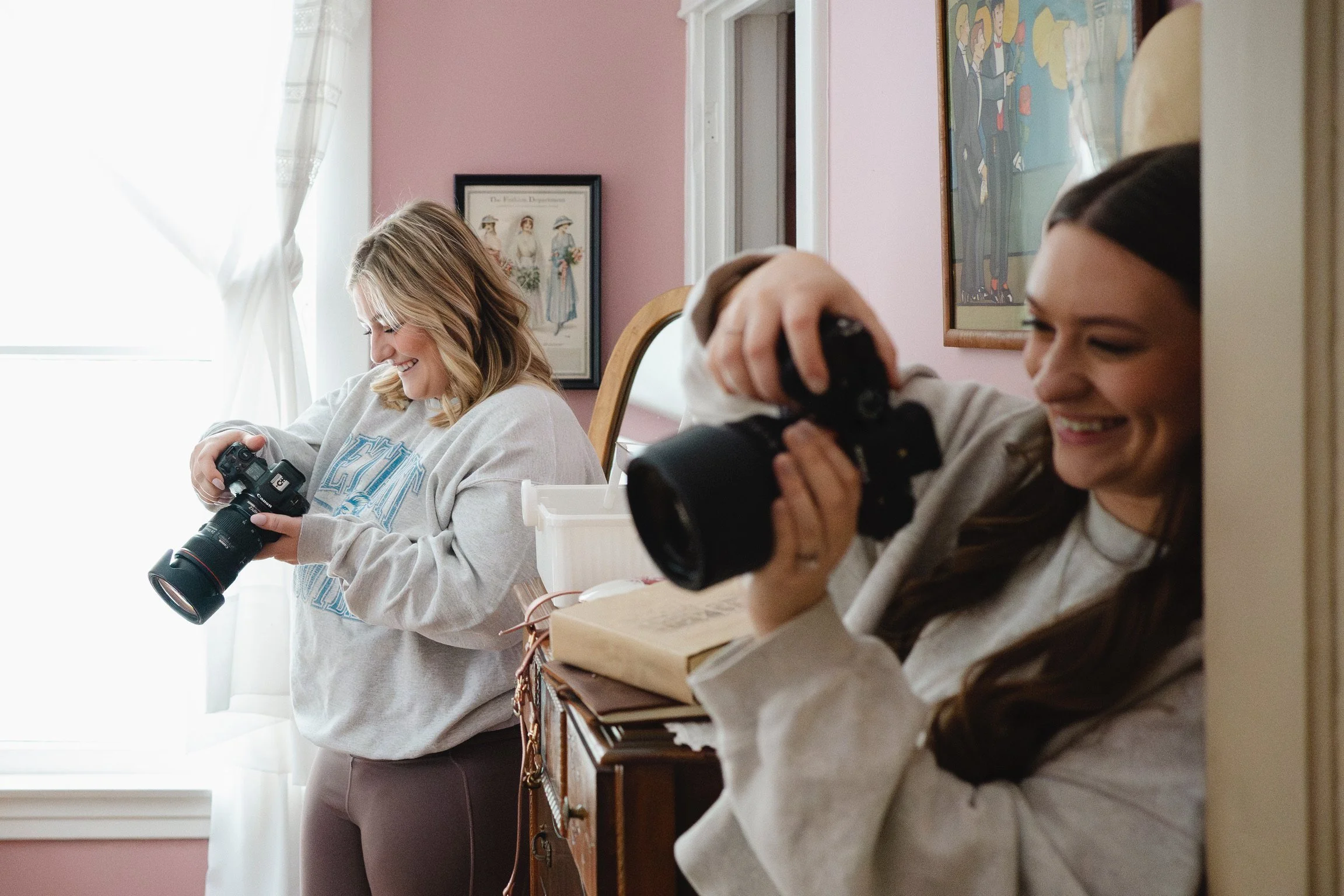 Two women are sitting at a table, one smiling and holding a camera, the other also smiling and looking at a camera. The room has pink walls and framed artwork hanging on the wall.