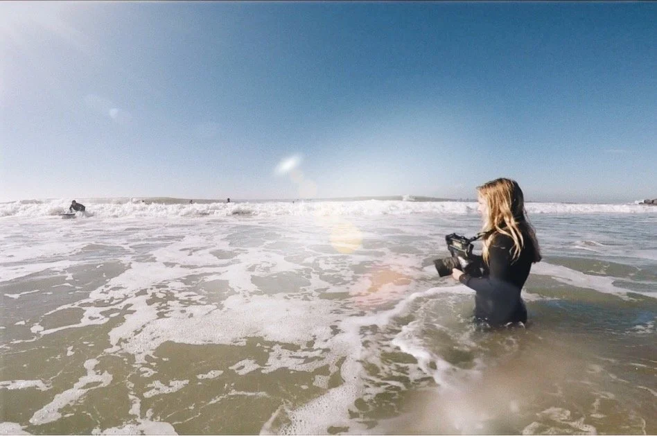 A woman with wavy blonde hair stands in the ocean holding a video camera, facing the horizon on a sunny day with a clear blue sky. There are waves and foam around her, and a few people are in the distance in the water.
