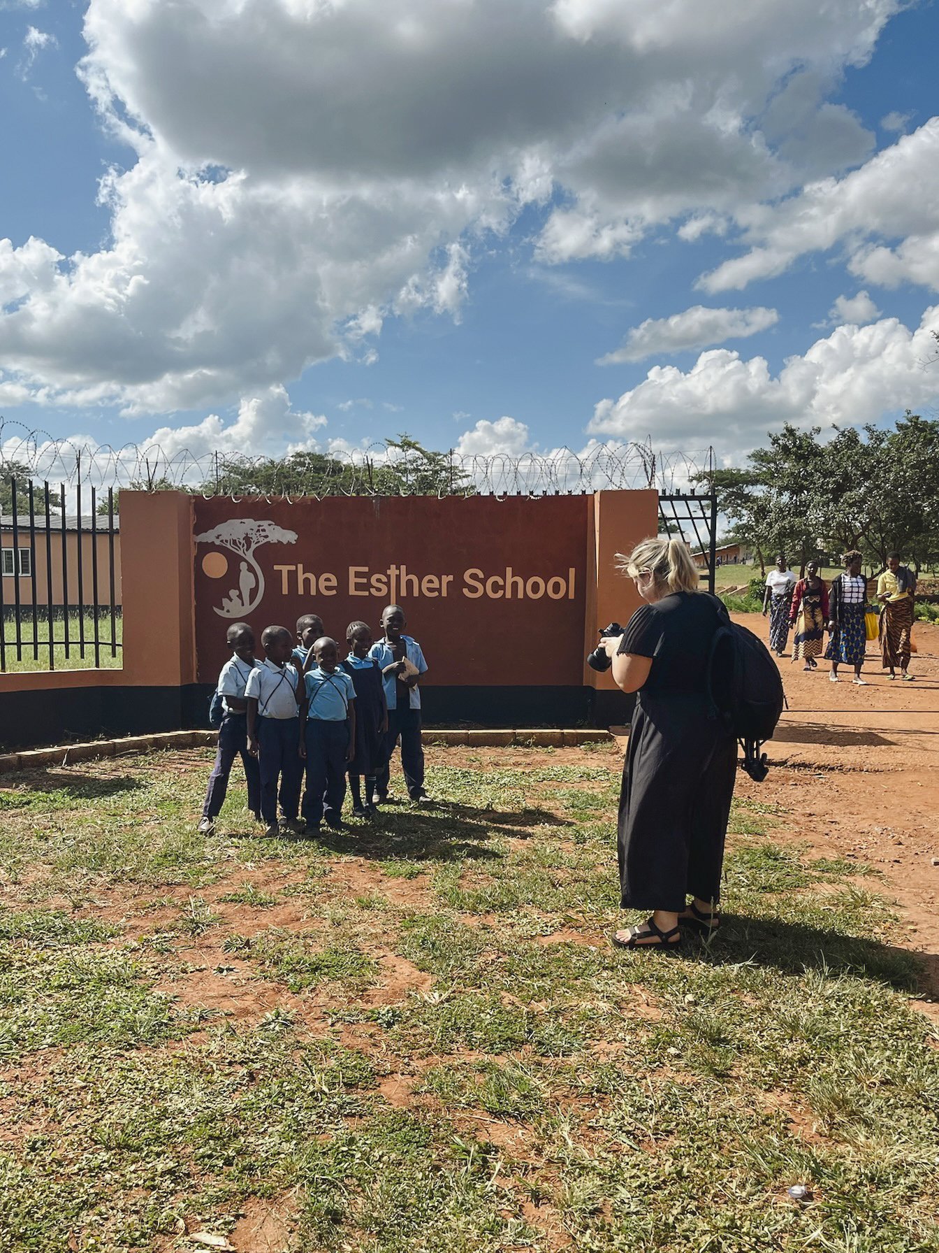 Group of school children and a woman with a camera in front of a school sign that reads "The Esther School", with a fence and trees in the background under a partly cloudy sky.